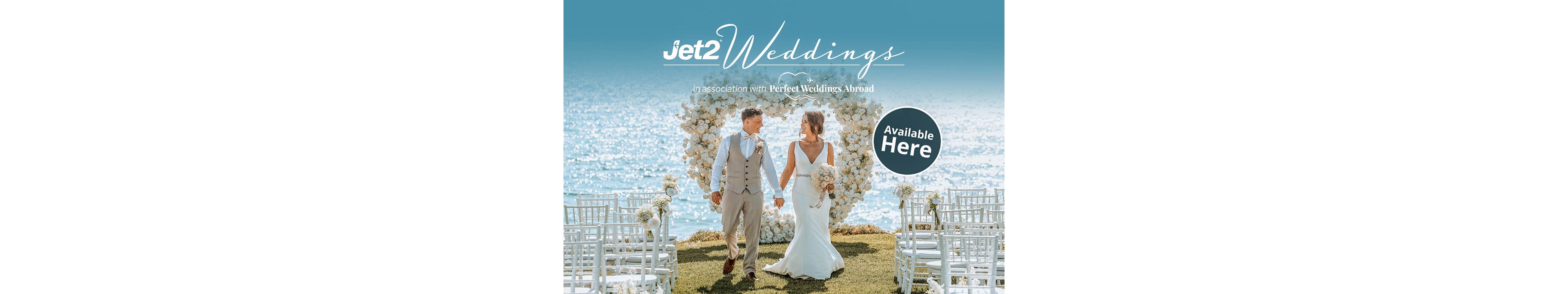 A bride and groom walk hand in hand down an outdoor aisle at a seaside wedding ceremony. Behind them is a large heart-shaped floral arrangement with the ocean sparkling in the background. White chairs and floral decorations line the grassy aisle, creating a romantic coastal setting. The text promotes Jet2 Weddings in association with Perfect Weddings Abroad, with a circular badge stating Available Here.
