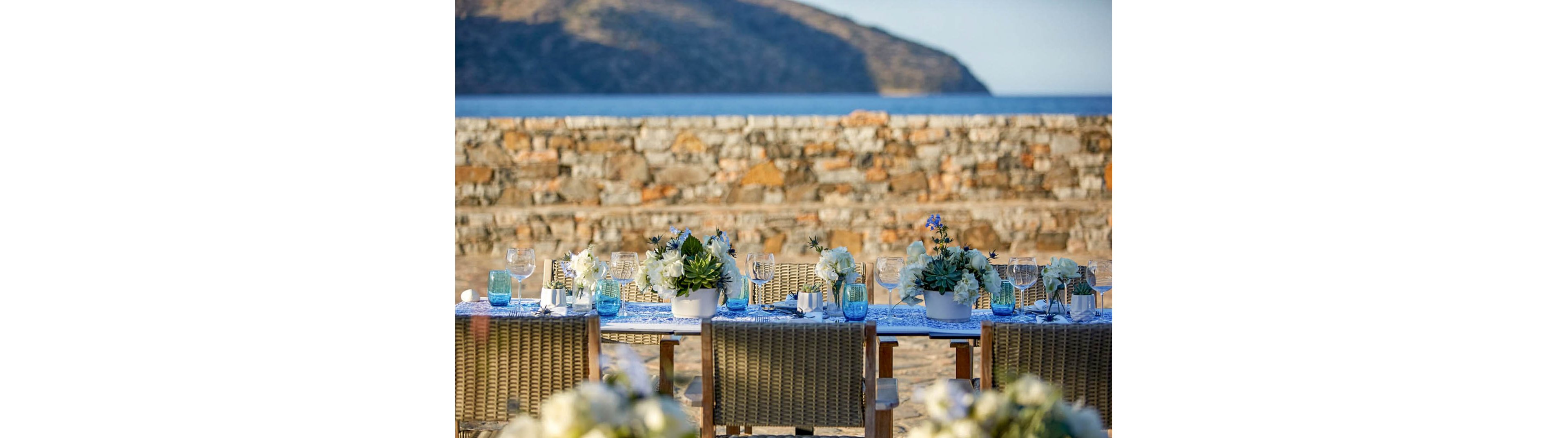 A long outdoor dining table is beautifully set with white floral arrangements and glassware. The table is positioned on a beachside terrace with woven chairs facing the sea. Blue glass vases and accents complement the coastal scenery and stone wall backdrop. Soft natural light enhances the tranquil, celebratory atmosphere by the water.
