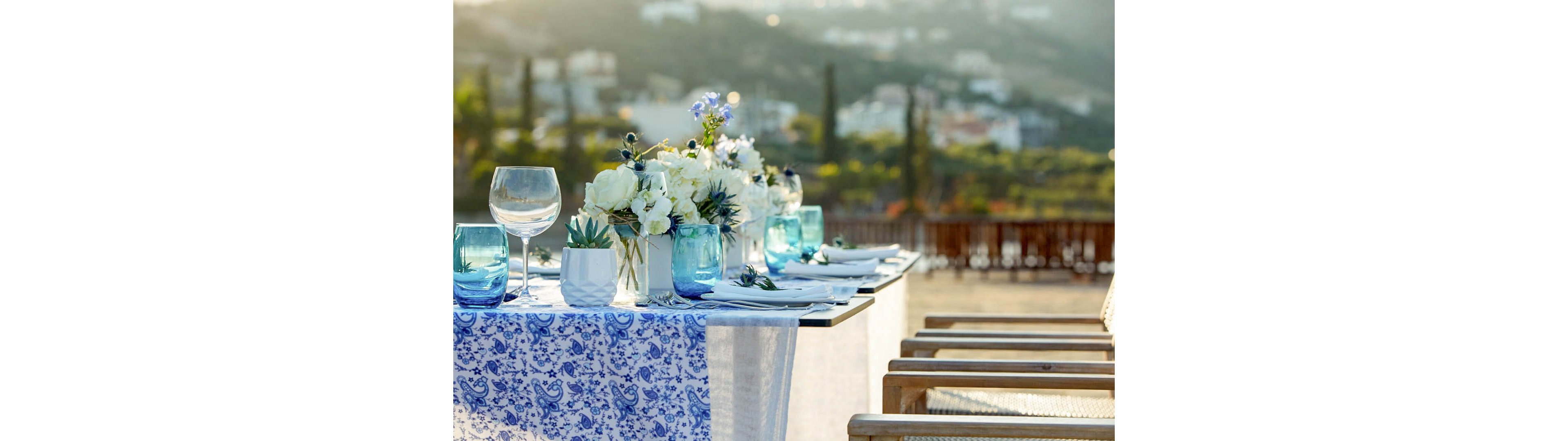 A beautifully arranged outdoor dining table is decorated with white flowers and greenery in simple vases. Blue-tinted glassware and a patterned blue and white table runner create a fresh coastal feel. Wooden chairs line the table, which is set with plates, napkins, and cutlery ready for guests. In the background, a sunlit hillside town and blurred landscape suggest a scenic terrace or patio setting.