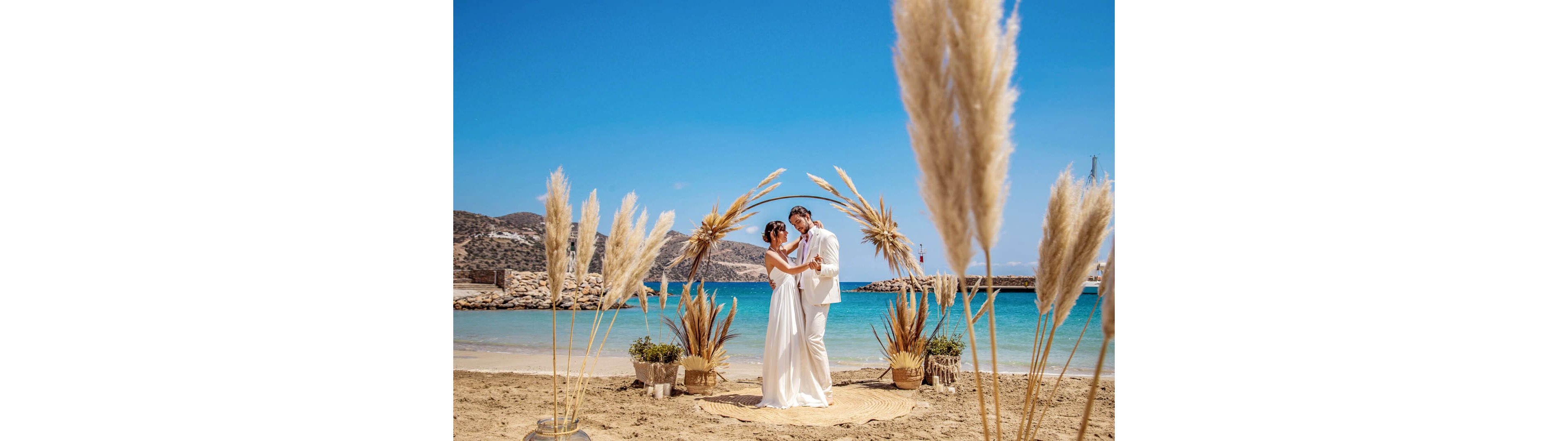 A newlywed couple stands embracing under a decorative arch on a sandy beach. The scene features pampas grass arrangements and rustic vases framing the couple in front of turquoise water. The groom wears a light suit while the bride wears a flowing white dress. Clear blue skies and a calm sea create a bright, romantic atmosphere.