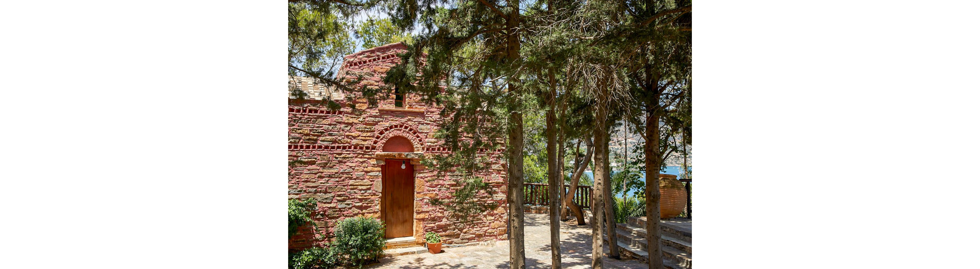 A small rustic chapel built from reddish stone stands in a shaded courtyard. The simple wooden door and arched brickwork give the building a historic, traditional character. Tall evergreen trees frame the scene, casting dappled shadows on the stone path and terrace. In the background, a railing and glimpse of blue water suggest a scenic hillside or coastal setting.