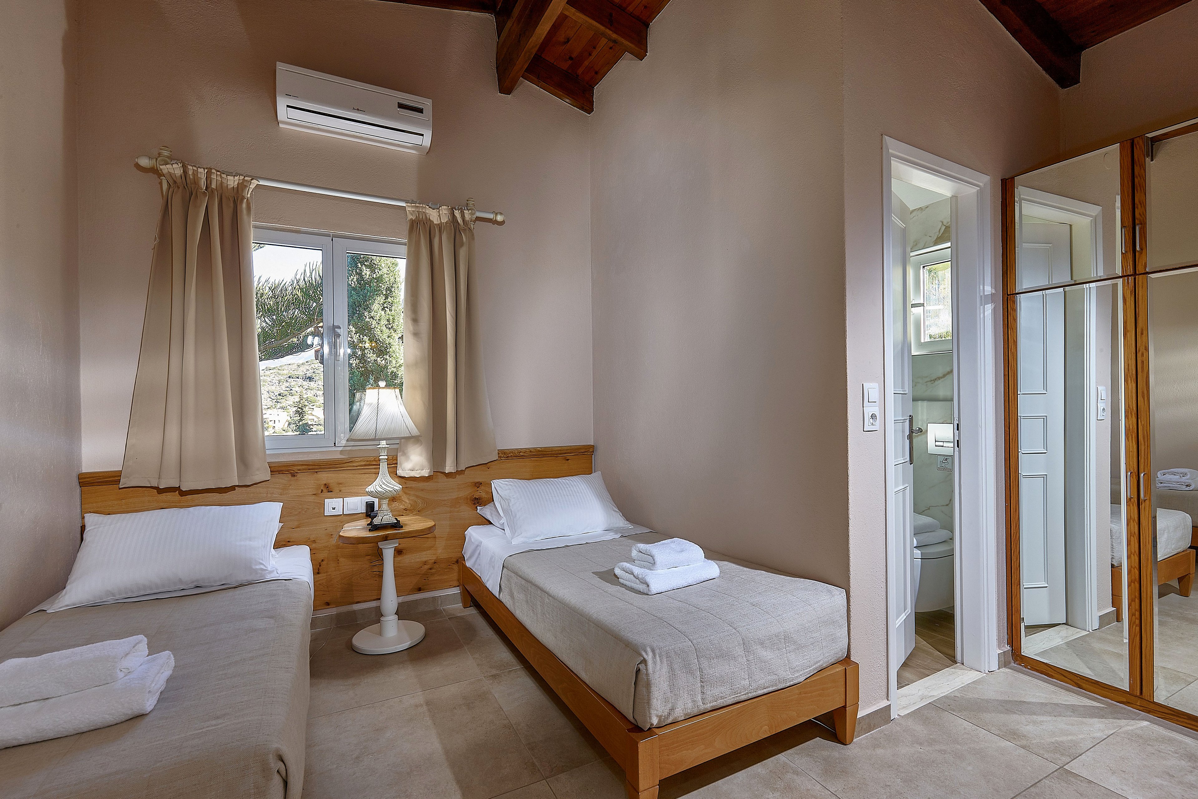 Bright twin bedroom featuring two single beds with neatly folded white towels. A small round bedside table with a lamp sits between the beds beneath a window with beige curtains. The room has tiled floors, light-colored walls, and a wooden ceiling, creating a warm and inviting atmosphere. A mirrored wardrobe reflects the interior, and an open door reveals a compact ensuite bathroom. An air conditioning unit is mounted above the window for added comfort.
