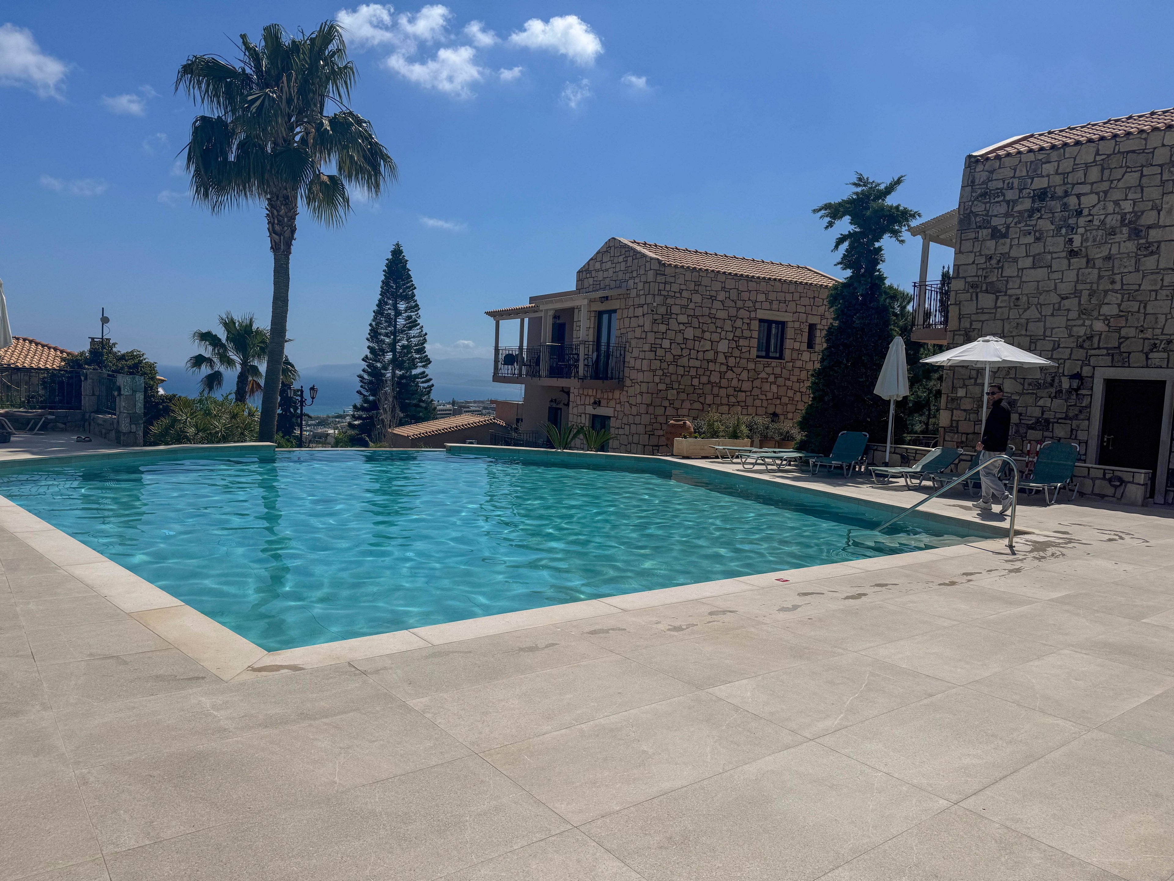 A clear blue swimming pool sits in the courtyard of a stone villa complex under a bright sunny sky. Lounge chairs and umbrellas line the poolside, suggesting a relaxing vacation setting. Palm trees and other greenery frame the scene, with glimpses of the sea visible in the distance. The overall atmosphere is calm, warm, and inviting, ideal for a resort or holiday stay.