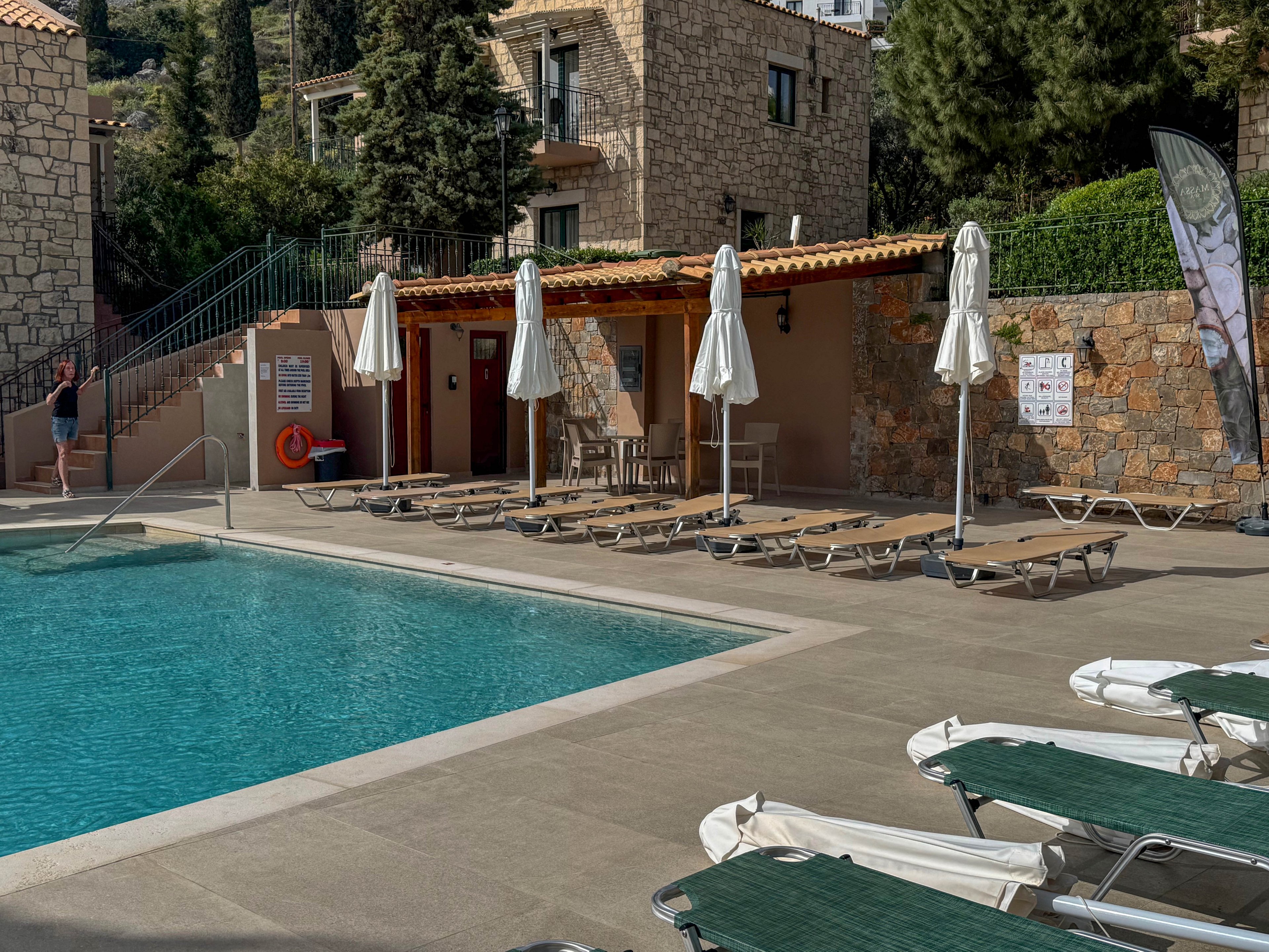 An outdoor hotel swimming pool is surrounded by empty sun loungers and closed white umbrellas. Stone buildings and lush greenery form the backdrop, creating a calm vacation setting. A single person stands near the stairs by the pool area, adding a sense of scale. The scene is brightly lit by daylight, emphasizing the clear blue water and warm stone textures.