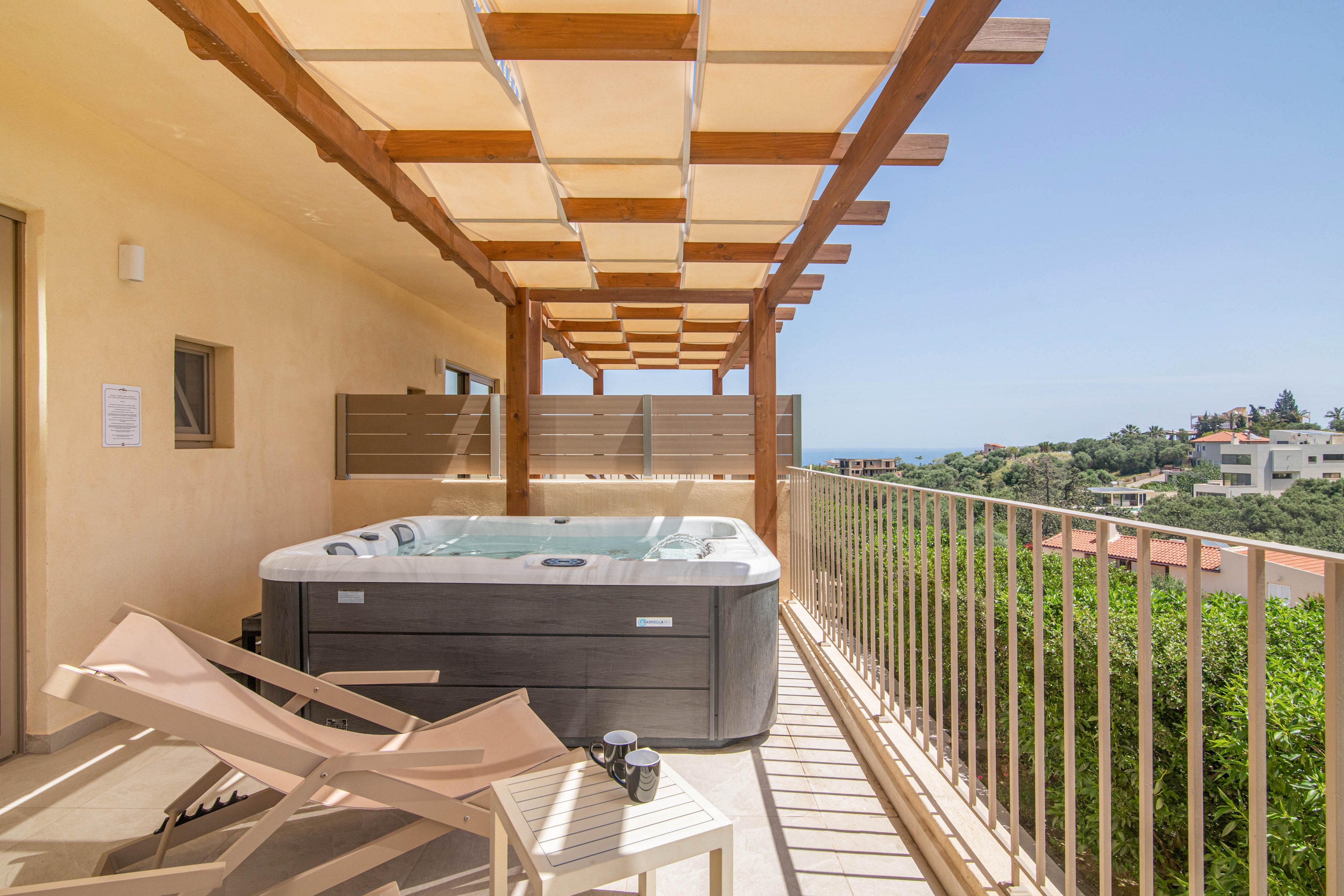 An outdoor terrace features a modern hot tub set against a light-colored wall. Two reclining lounge chairs and a small side table with cups create a relaxing seating area. A wooden pergola with geometric panels provides partial shade above the deck. The terrace overlooks lush greenery and distant buildings under a clear blue sky, suggesting a serene vacation setting.