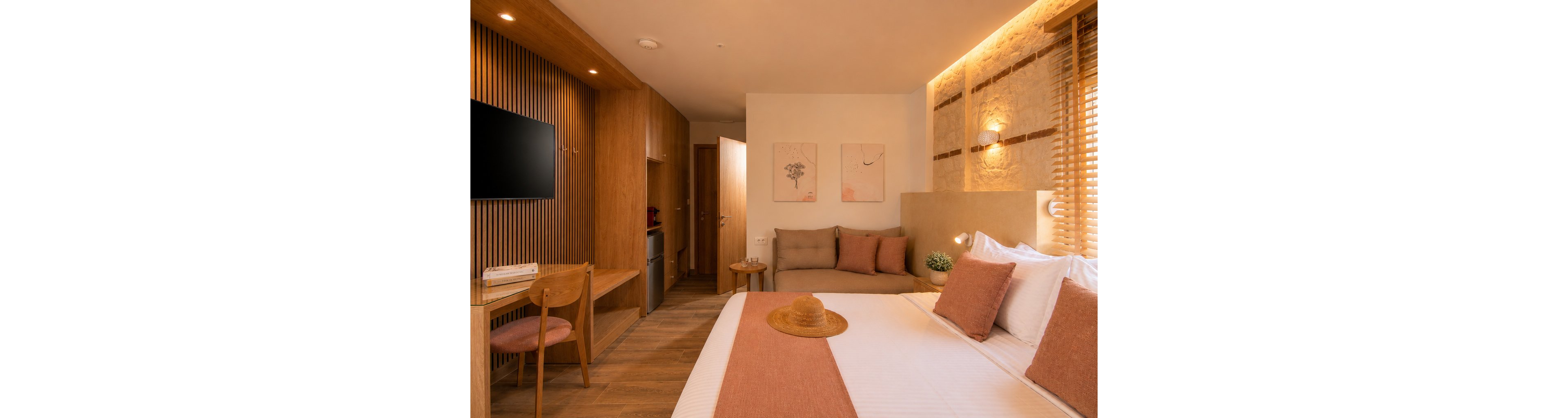 A cozy hotel room features a neatly made bed with soft pillows and a straw hat placed on top. The space combines wooden furniture, including a desk, chair, and built-in cabinetry, with warm neutral textiles in beige and terracotta tones. A wall-mounted television, small sofa, and decorative artwork add comfort and style to the compact room. Soft lighting from recessed ceiling lights and wall sconces creates a welcoming, relaxing atmosphere.