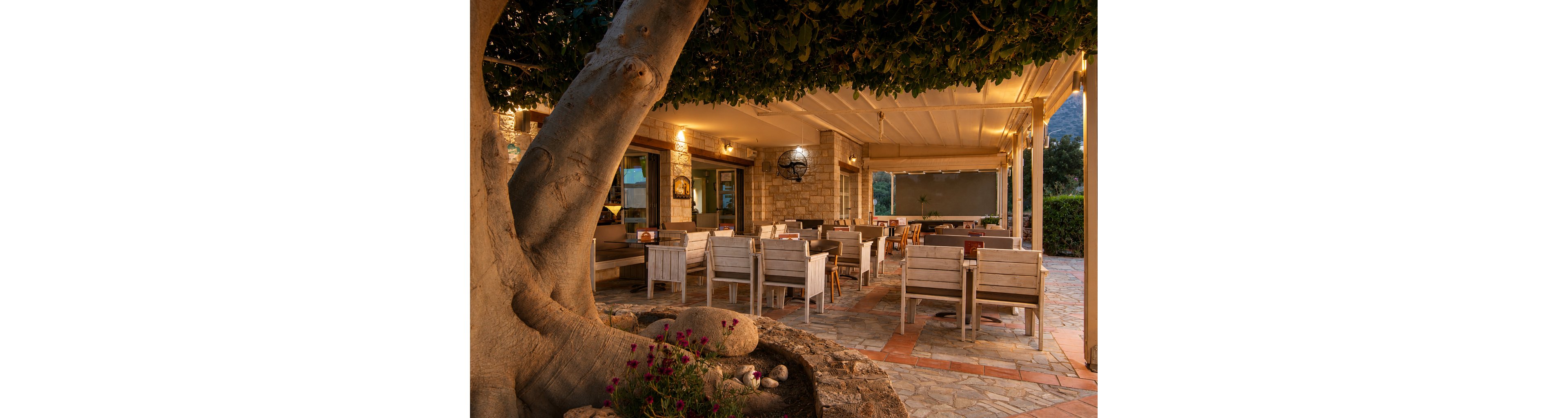 An inviting outdoor restaurant terrace features wooden tables and chairs arranged beneath a covered patio. Warm ambient lighting glows from the ceiling and interior windows, creating a cozy evening atmosphere. A large tree trunk and stone planter frame the foreground, adding natural texture to the stone-paved floor. The setting appears calm and empty, ready to welcome guests for a relaxed dining experience.