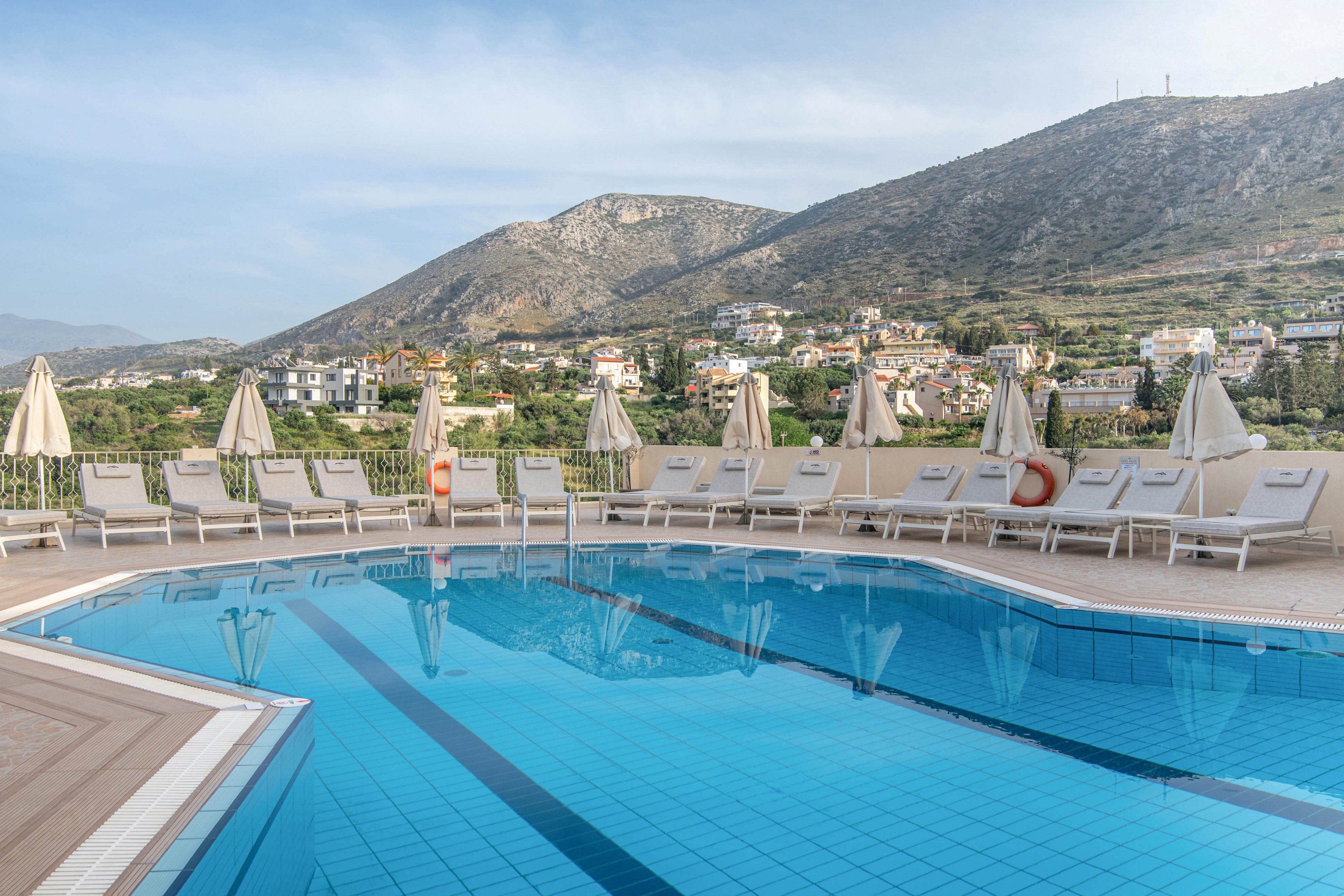 An outdoor hotel swimming pool sits in the foreground, reflecting the clear sky and surrounding umbrellas. Numerous sun loungers and closed parasols line the poolside terrace, suggesting a quiet, early morning or off-peak moment. In the background, a hillside town with scattered houses rises up the slopes of a large mountain. The overall scene feels calm and inviting, with soft natural light and a relaxed resort atmosphere.
