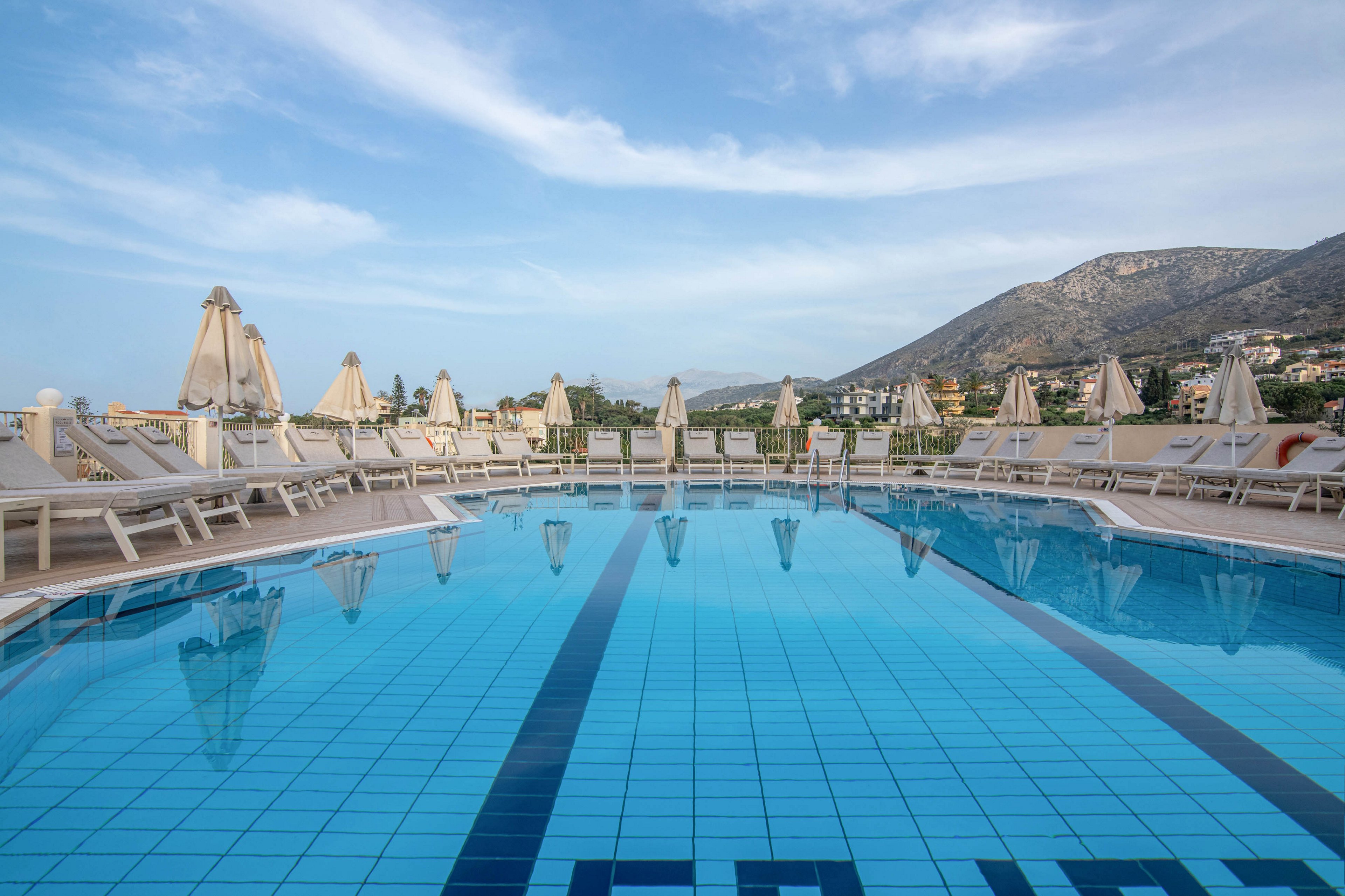 A clear blue swimming pool is surrounded by rows of white lounge chairs and closed umbrellas. The scene appears to be an empty resort terrace overlooking a town. In the background, rolling hills and mountains rise under a soft, partly cloudy sky. The calm water reflects the loungers and umbrellas, creating a tranquil vacation atmosphere.