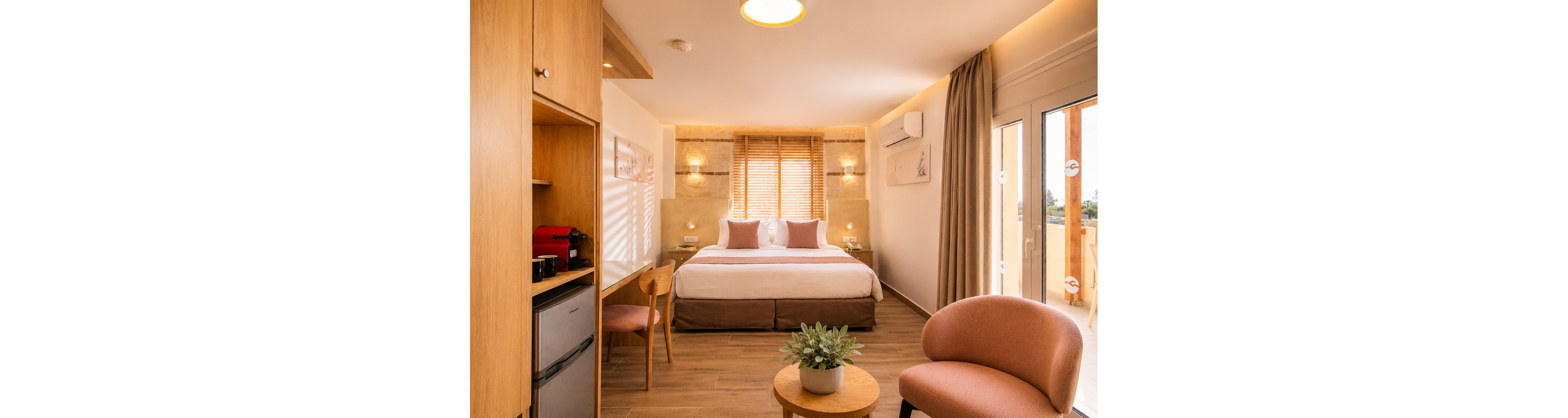 A contemporary hotel bedroom features a large double bed with plush pillows and neutral linens. Warm wooden furniture, including a desk, chair, and small round table with a potted plant, creates a cozy atmosphere. A compact fridge and coffee machine are built into a wooden cabinet on the left side. Large windows with curtains and glass doors open to a balcony, allowing natural light to fill the room. Soft beige and blush tones enhance the inviting, minimalist design.