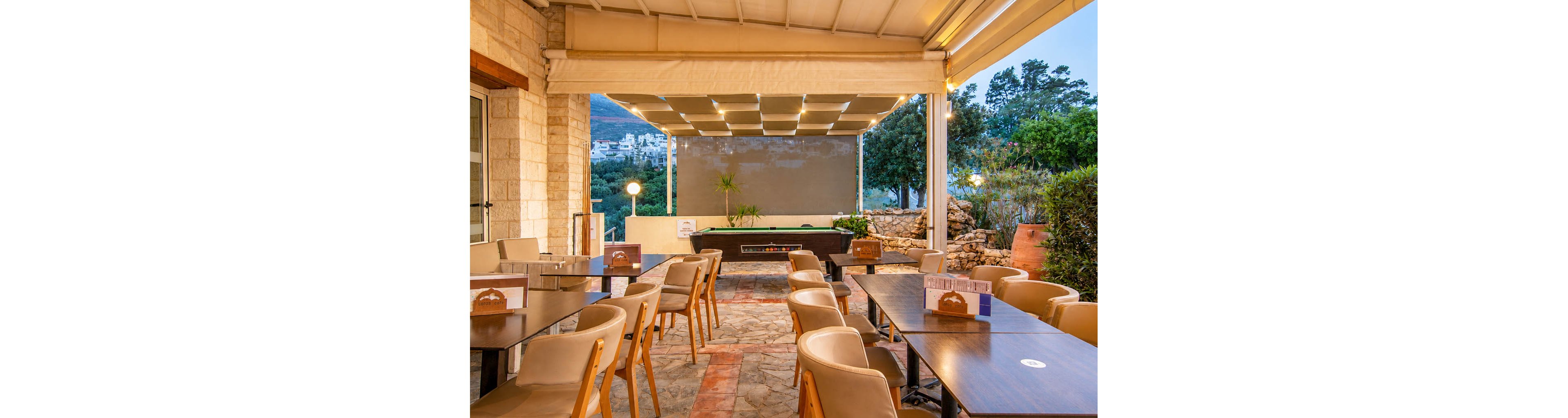 An open-air restaurant terrace features neatly arranged tables and chairs under a covered pergola. The stone floor and warm-toned furniture create a welcoming dining area, currently empty of guests. In the background, greenery and distant buildings on a hillside add depth to the scene. Soft evening lighting enhances the relaxed, tranquil atmosphere of the outdoor space.