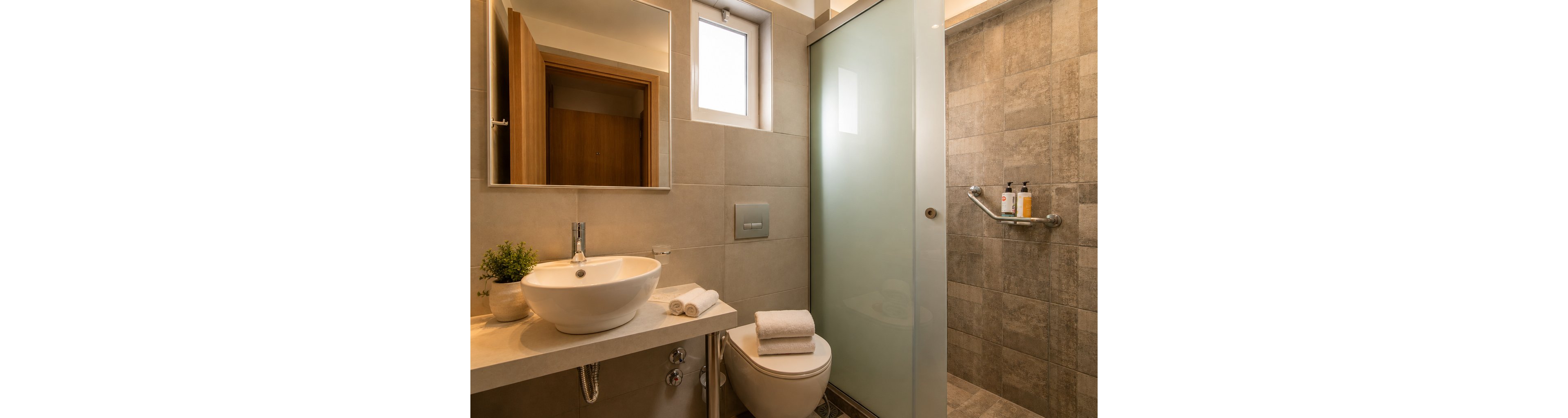 A contemporary bathroom features a round vessel sink on a light countertop beside a compact toilet. A frosted glass door separates the vanity area from a tiled walk-in shower. Soft beige and gray tiles create a warm, neutral palette throughout the space. Rolled white towels and a small potted plant add spa-like accents, while toiletries rest on a metal shower rail.