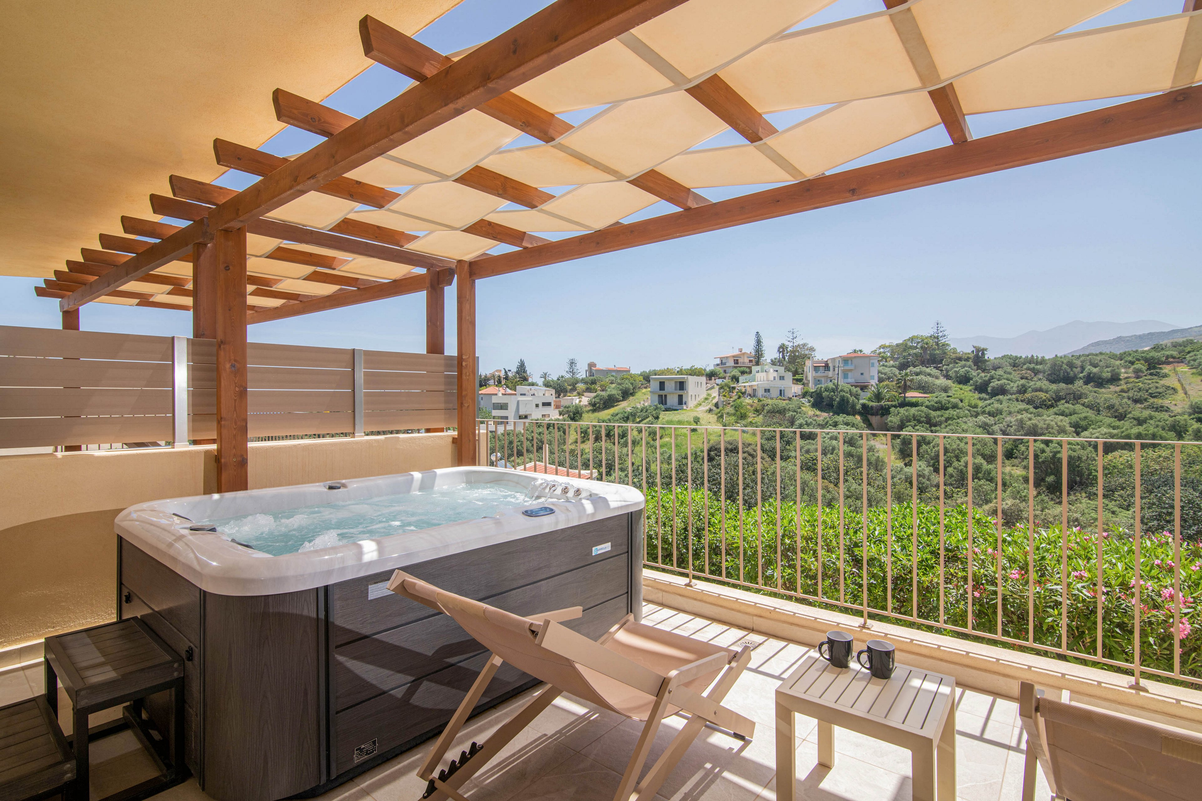 An outdoor terrace features a modern hot tub under a wooden pergola. Two reclining deck chairs and a small table with cups create a relaxed seating area. Beyond the railing, a lush green hillside and scattered houses stretch into the distance under a clear sky. The bright daylight and neutral tones give the scene a calm, vacation-like atmosphere.