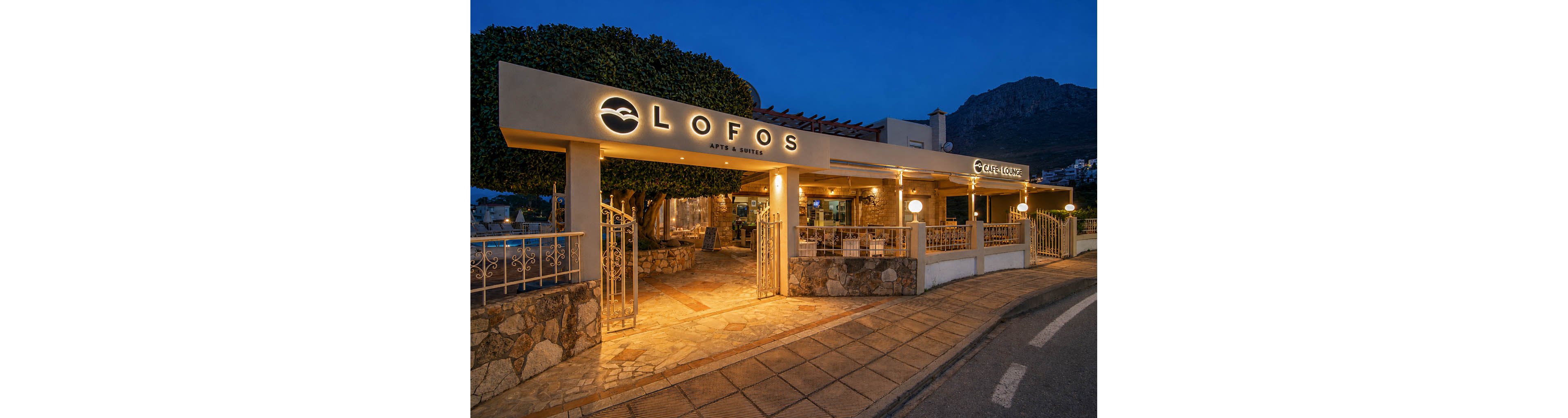 An illuminated entrance to Lofos Apts & Suites is shown at blue hour, with warm lights glowing against the evening sky. The exterior features stone walls, metal gates, and a covered terrace area. A sign for a café lounge is visible inside the open-air structure. The scene captures a quiet, inviting atmosphere along a gently curving street with mountains in the background.