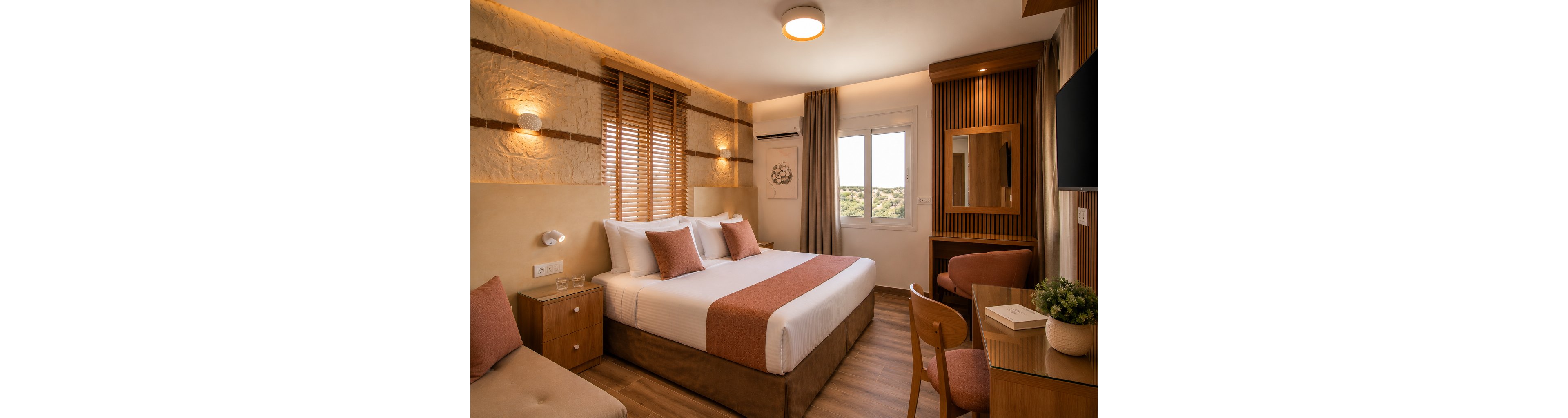 A neatly made double bed with white linens and muted terracotta accents is the focal point of this cozy hotel room. The space features warm wooden flooring, a small seating area with a chair and table, and a built-in desk with a mirror. Natural light enters through a window with wooden blinds, complemented by soft wall and ceiling lighting. Neutral walls with textured stone details and coordinated textiles create a warm, inviting atmosphere.