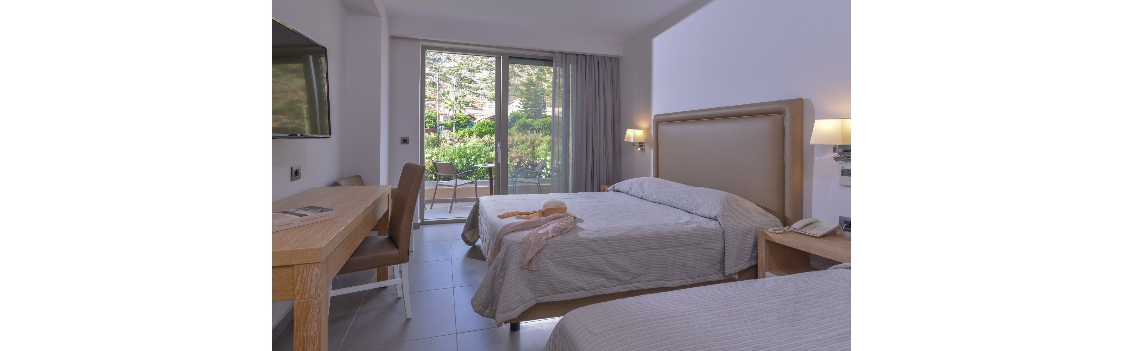 A contemporary hotel room features two twin beds with neutral bedding, a wooden desk, and a wall-mounted television. The space is bright, with natural light streaming in from a glass door leading to a private patio overlooking lush greenery. The room is tidy, with a hat and scarf placed on one bed, creating a relaxed and inviting atmosphere.