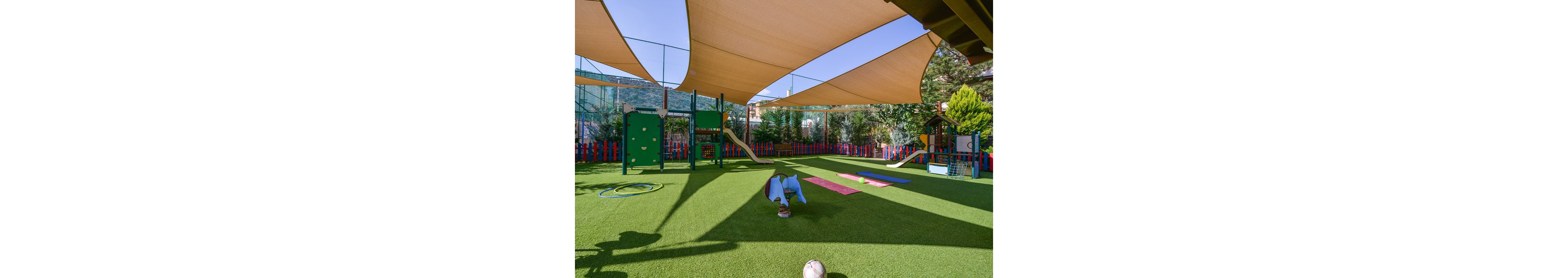 A vibrant outdoor playground features artificial grass, climbing equipment, and slides under large beige sun shades. The area is surrounded by a colorful fence and includes play mats, a toy elephant rocker, and a soccer ball. The setting is bright and cheerful, designed for young children’s activities and play.
