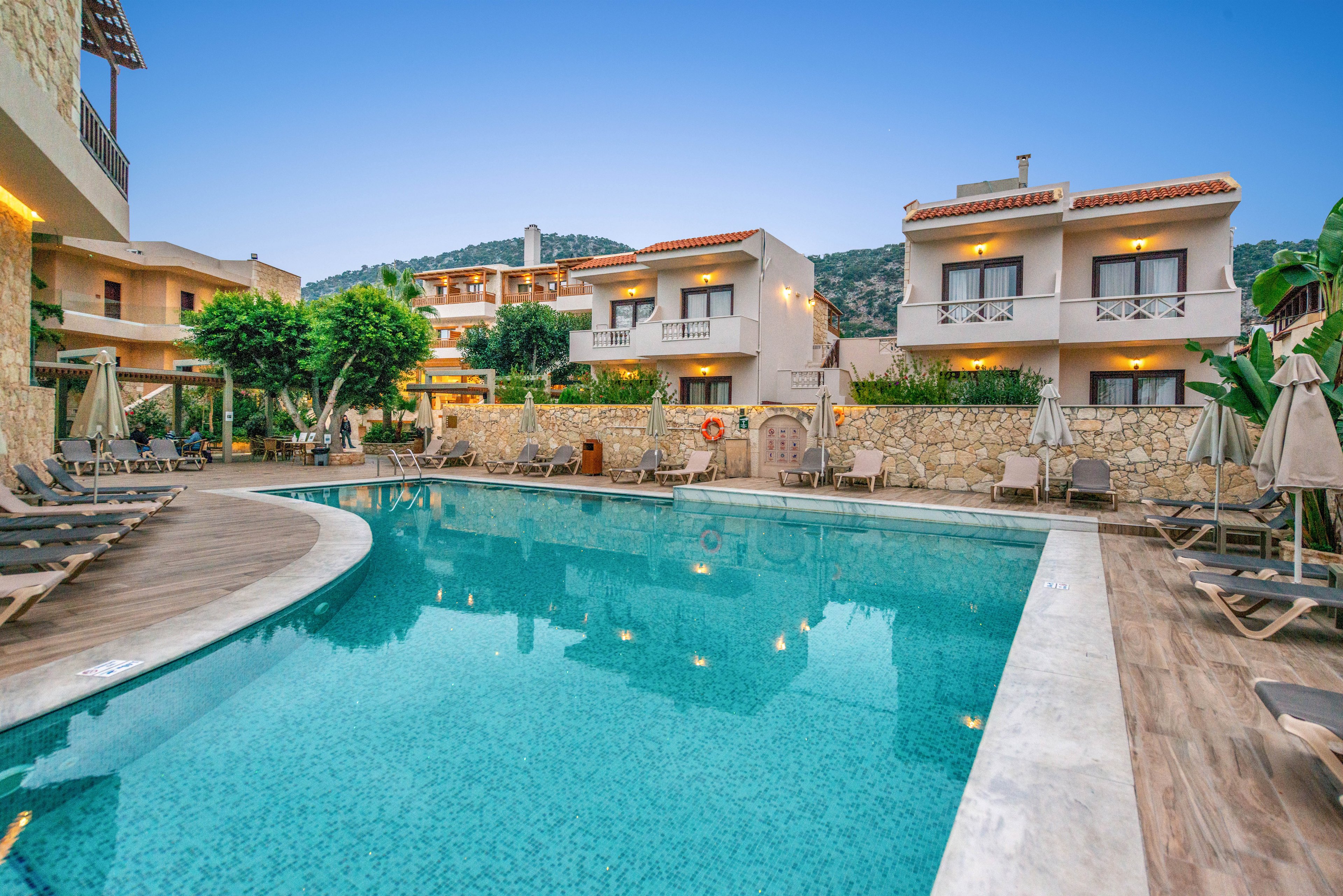 A serene outdoor swimming pool is surrounded by modern resort buildings and sun loungers. The setting is tranquil, with clear blue water reflecting the evening sky and nearby architecture. The area is well-maintained, featuring stone walls, lush greenery, and a mountain backdrop. No visible people or text are present in the image.