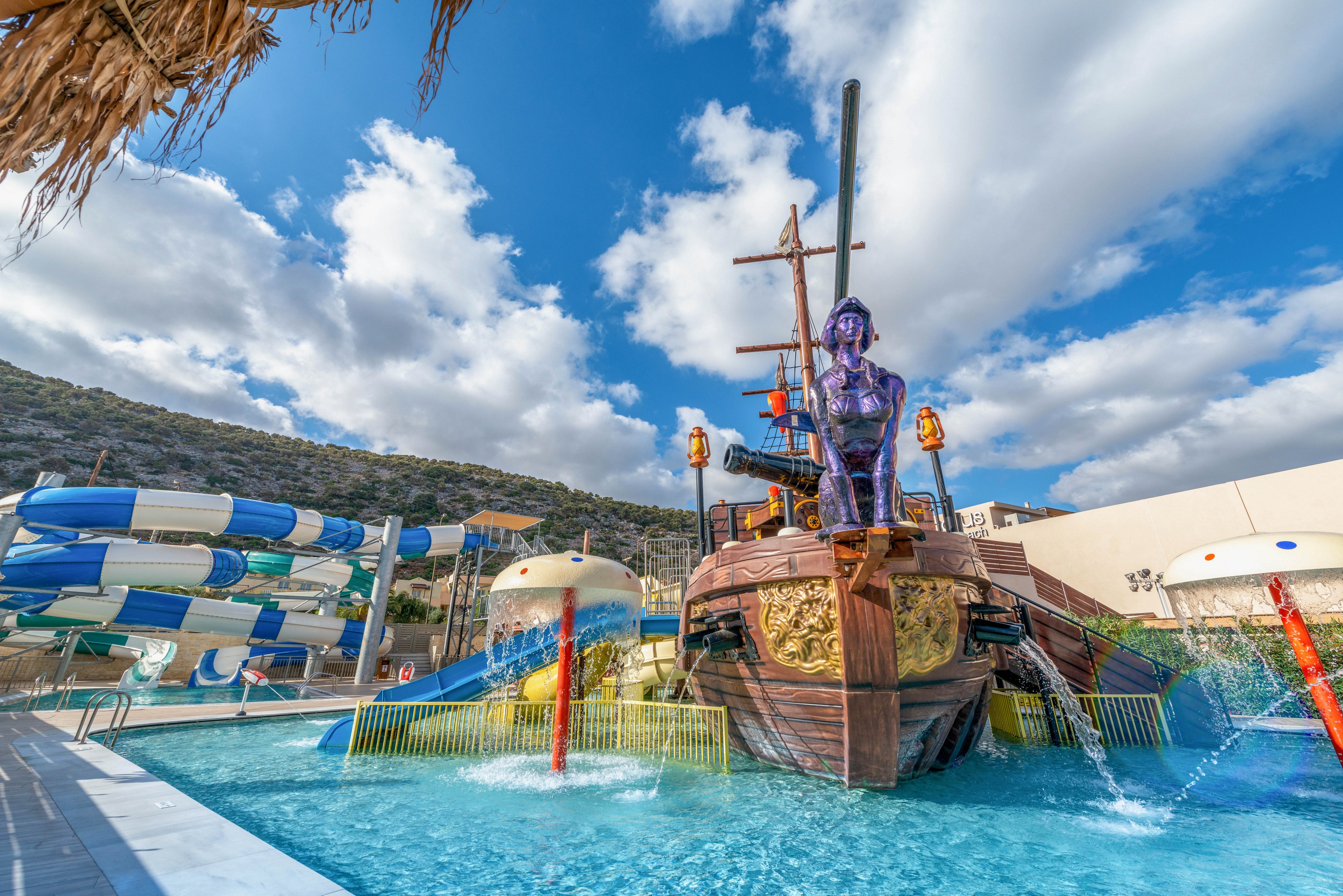 A large pirate ship-themed water play structure is the main feature in this outdoor water park. The setting includes blue and white water slides, splash zones, and mushroom-shaped fountains, all surrounded by hills and partly cloudy skies. The visuals are vibrant and playful, with bright colors and whimsical design elements. No visible text or numeric values are present in the image.