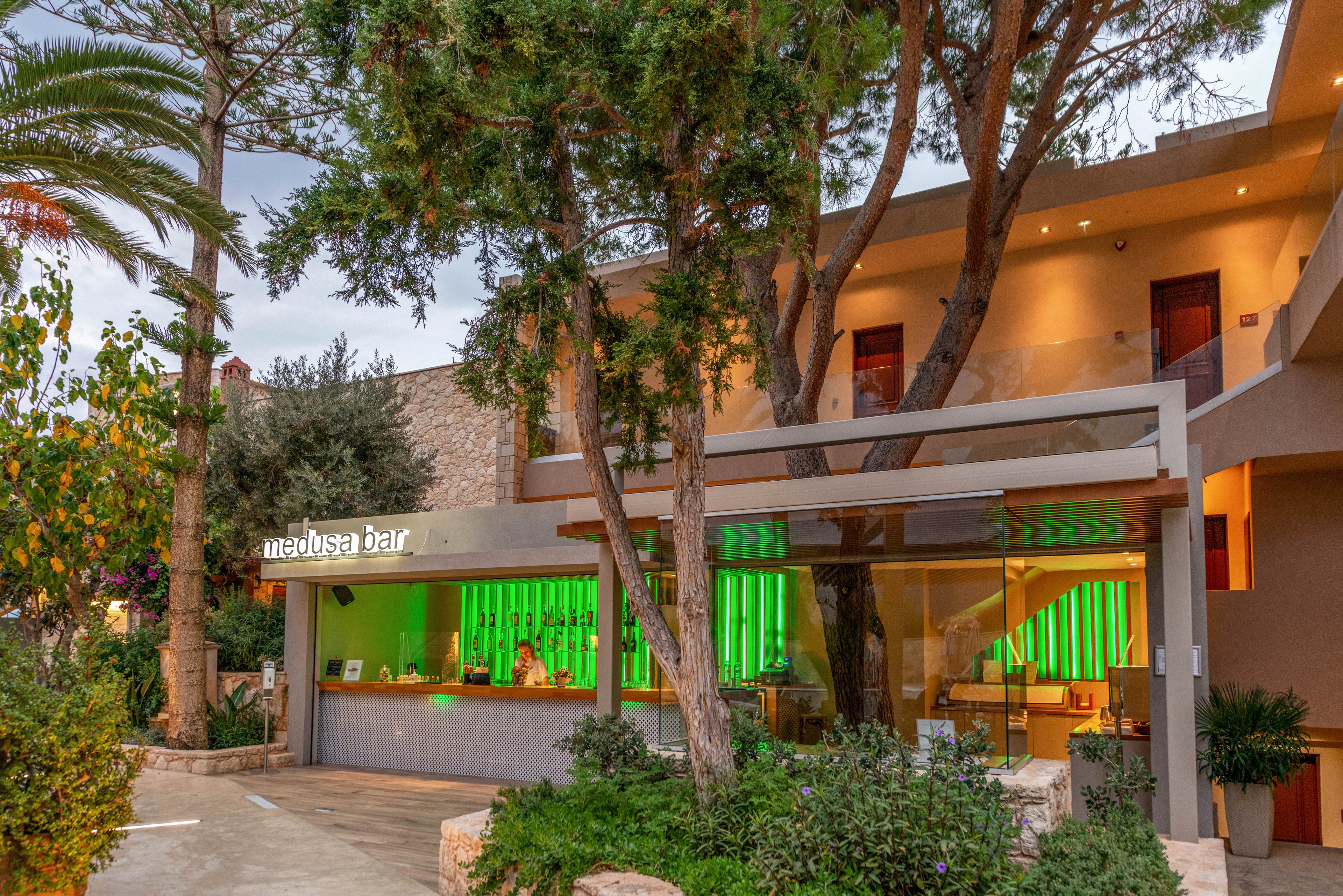 The image shows the exterior of the Medusa Bar, featuring a contemporary architectural design with a prominent green-lit bar area. The setting is outdoors, surrounded by trees and lush greenery, creating a relaxed and inviting atmosphere. The bar's name 'medusa bar' is clearly visible in illuminated signage. The overall mood is vibrant and welcoming, with warm evening lighting and natural elements.