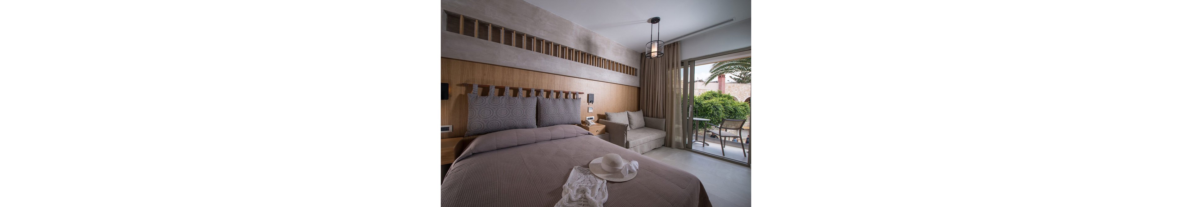 A contemporary hotel room features a neatly made double bed with patterned pillows and a taupe bedspread. The room includes a small sofa, wooden accents, and a sliding glass door leading to a balcony with outdoor seating and lush greenery. Soft natural light fills the space, and a white sunhat and lace garment rest on the bed, suggesting a relaxed, vacation mood.