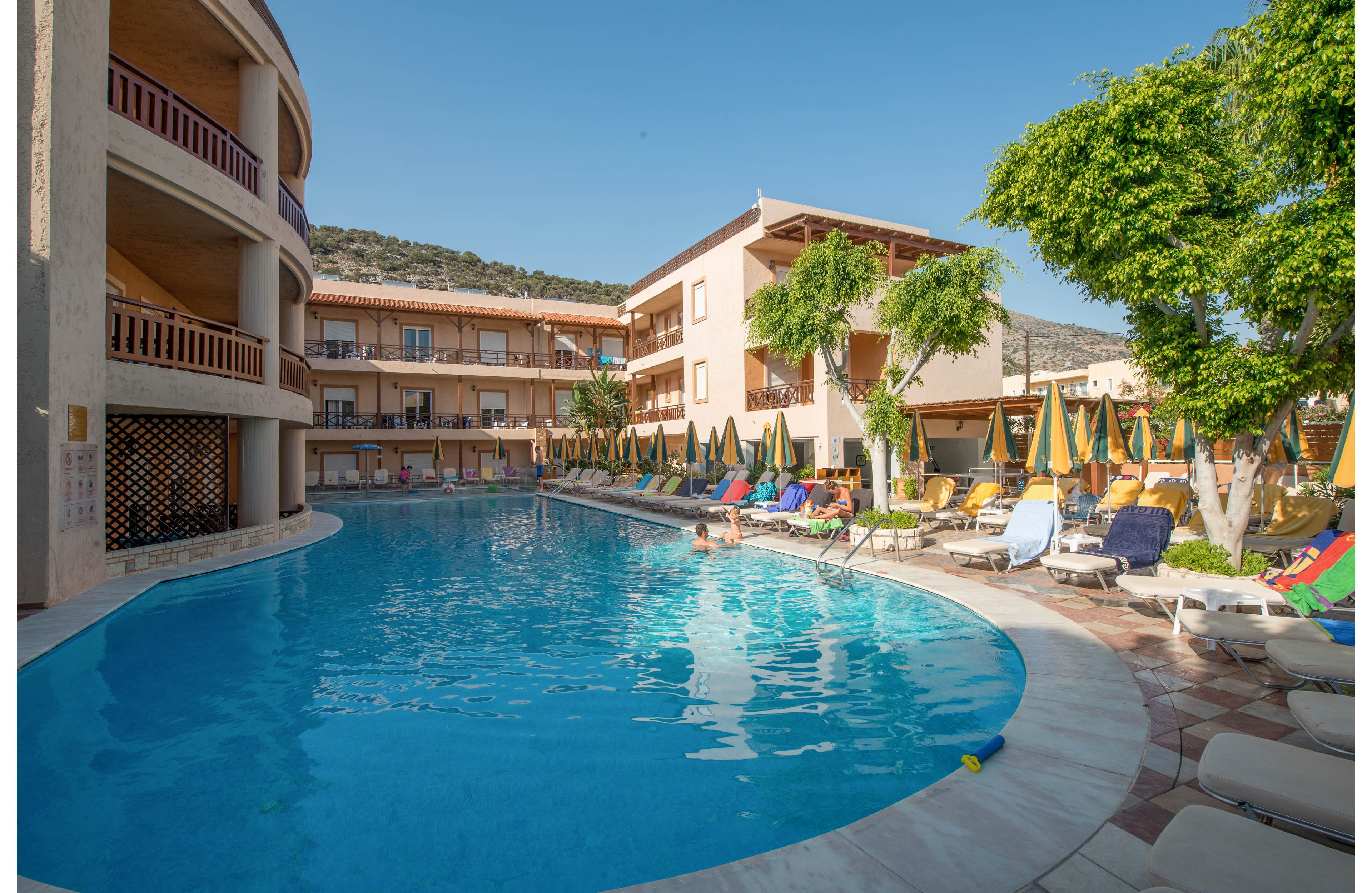 A large, curved outdoor swimming pool is surrounded by sun loungers and umbrellas at a resort hotel. The setting is bright and sunny, with clear blue skies and modern, beige-colored buildings in the background. Several people are relaxing by the poolside, enjoying the leisure atmosphere. Green trees and mountain scenery add to the tranquil vacation vibe. The overall mood is relaxed and inviting, ideal for holidaymakers.