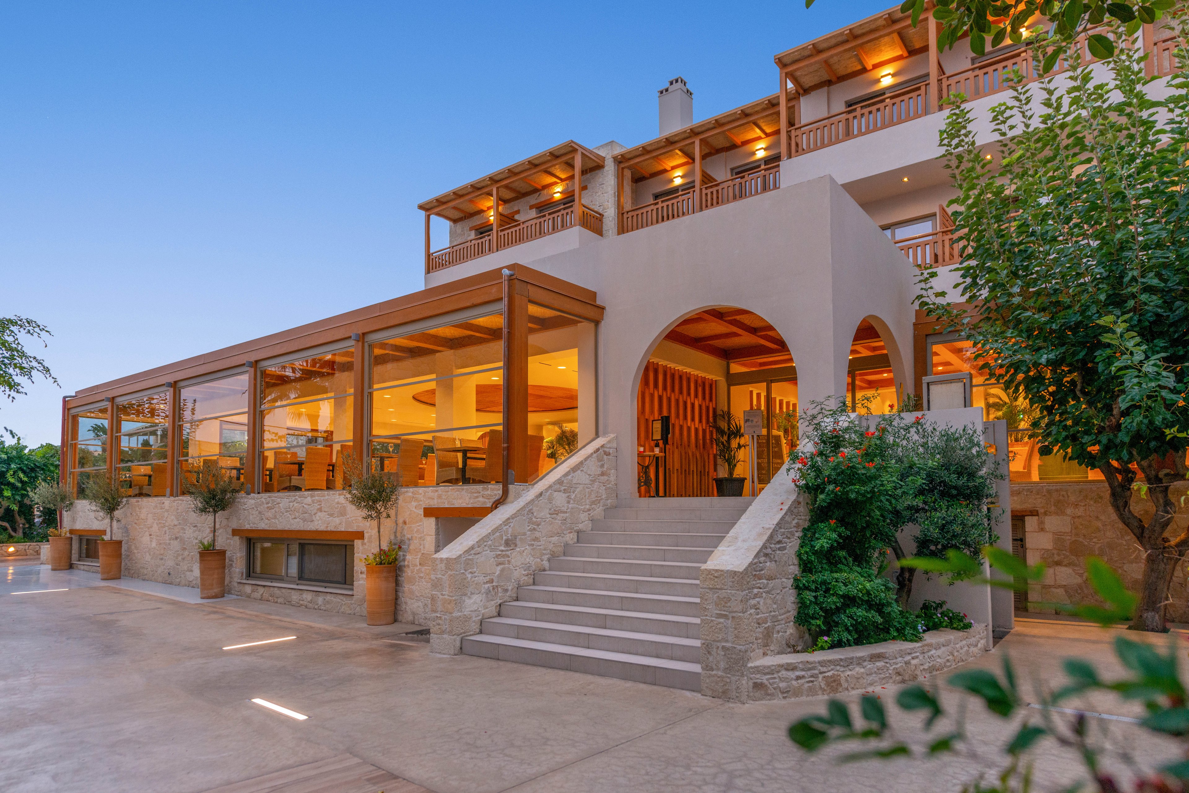 A contemporary hotel building features a spacious outdoor terrace and large windows, warmly illuminated at dusk. The setting includes stone steps, potted plants, and wooden architectural accents, creating a welcoming atmosphere. The sky is clear, and the overall mood is tranquil and inviting, ideal for hospitality or travel themes.