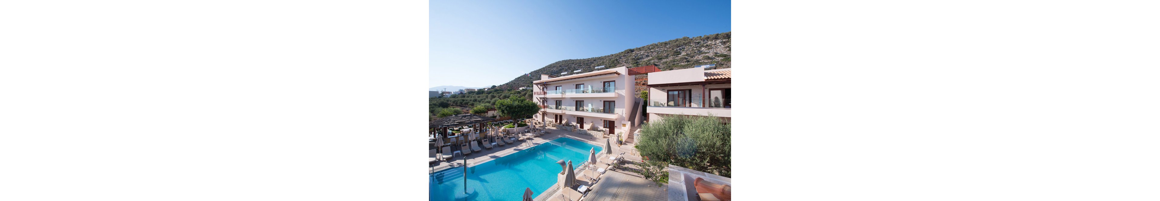 A contemporary hotel complex features a spacious outdoor swimming pool surrounded by lounge chairs and umbrellas. The setting is sunny and tranquil, with the hotel nestled against a scenic hillside. The architecture is clean and modern, with light-colored facades and balconies overlooking the pool area. No visible text or numeric values are present in the image.