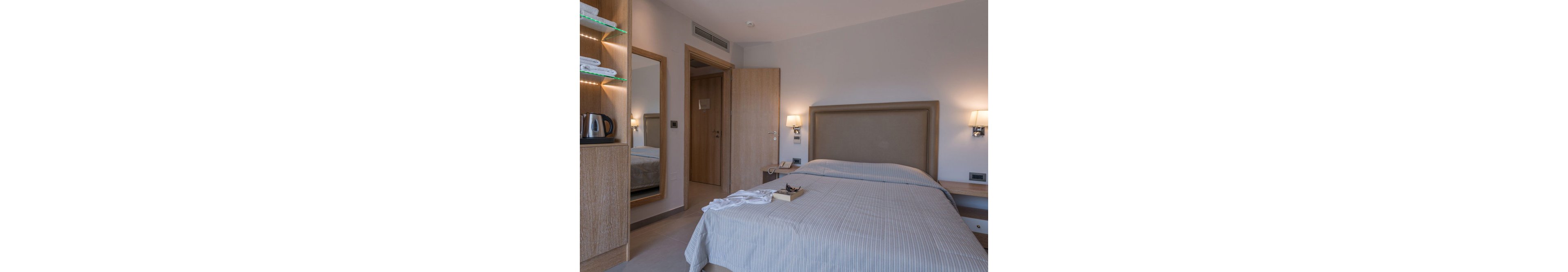 A contemporary hotel room features a neatly made bed with striped linens, wooden headboard, and bedside lamps. The setting includes a wall mirror, open shelving with towels, and a kettle visible on the shelf. The room is illuminated with soft lighting, creating a calm and inviting atmosphere. No visible numeric values or identifiable brands are present.