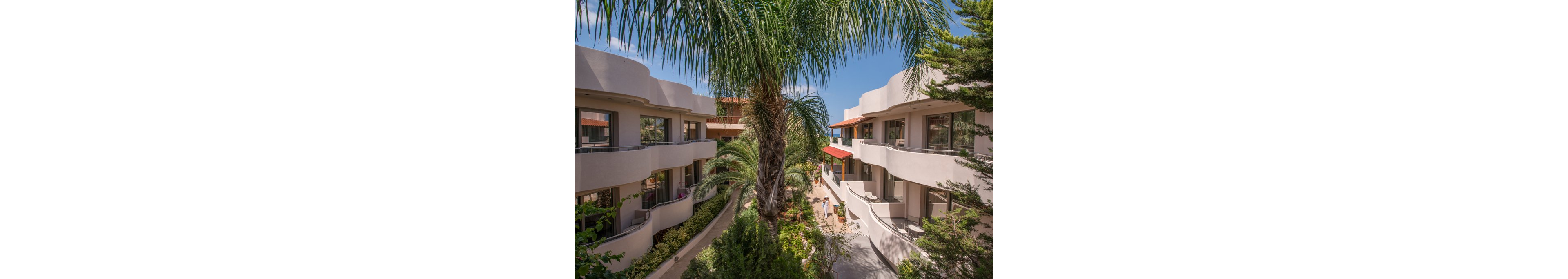The image features contemporary resort-style buildings with curved balconies and large windows, surrounded by vibrant palm trees and greenery. The setting appears to be a sunny, tropical or Mediterranean location, with clear blue skies overhead. The architectural style is modern, and the mood is relaxed and inviting, ideal for vacation or leisure. No visible text or numeric values are present in the image.