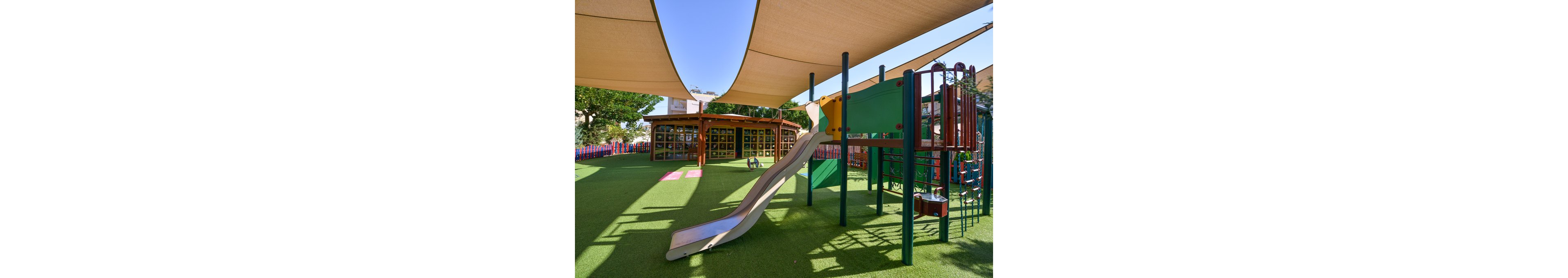 A contemporary playground features a slide, climbing structures, and artificial turf under large shade sails. The area is empty, with no children present, and is surrounded by colorful fencing and a building with large windows. The scene is brightly lit, suggesting a sunny day, and the overall atmosphere is clean and inviting.