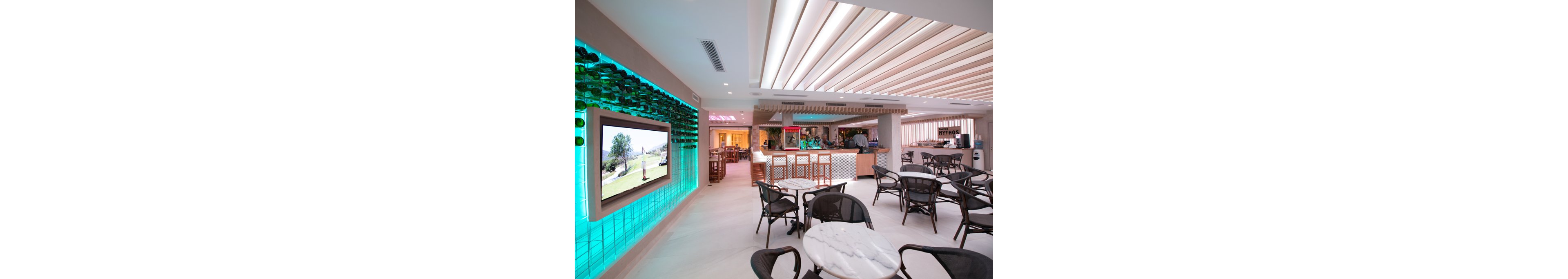 A contemporary café interior features marble-topped tables and black chairs arranged in a spacious, well-lit setting. The space is accented by a wall with green glass bottles and a large digital screen displaying a landscape. The ceiling has geometric lighting, and the bar area is visible in the background with a sign reading 'MYTHOS.' The overall palette is light with teal and pink neon highlights.