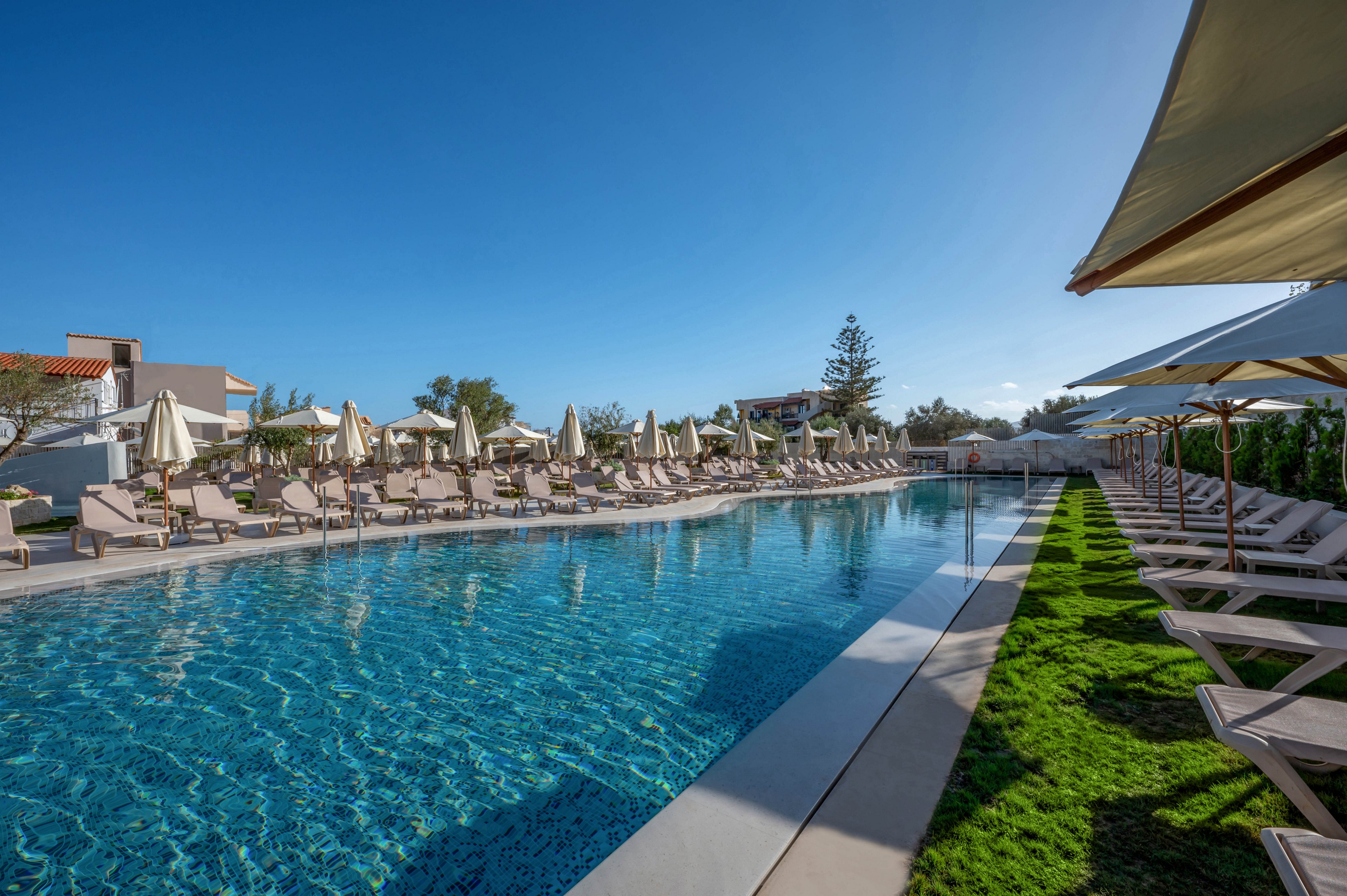 A large outdoor swimming pool is surrounded by numerous beige sun loungers and umbrellas, set in a modern hotel or resort environment. The scene is bathed in bright sunlight, with clear blue skies and lush green grass bordering the pool area. No people are present, creating a tranquil and inviting atmosphere for relaxation. The pool water is sparkling and reflects the surrounding architecture.