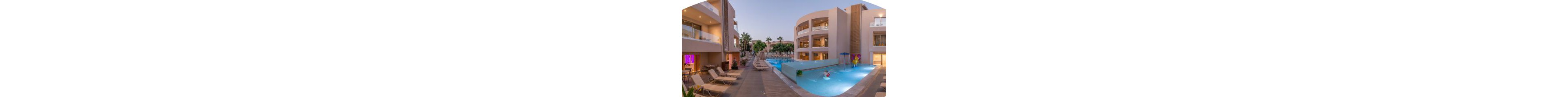 A contemporary resort features a central swimming pool surrounded by multi-story buildings with balconies. The setting is tranquil at dusk, with lounge chairs arranged poolside and palm trees visible in the background. The pool includes playful water features and a shallow area for children, while the overall palette is soft and inviting.