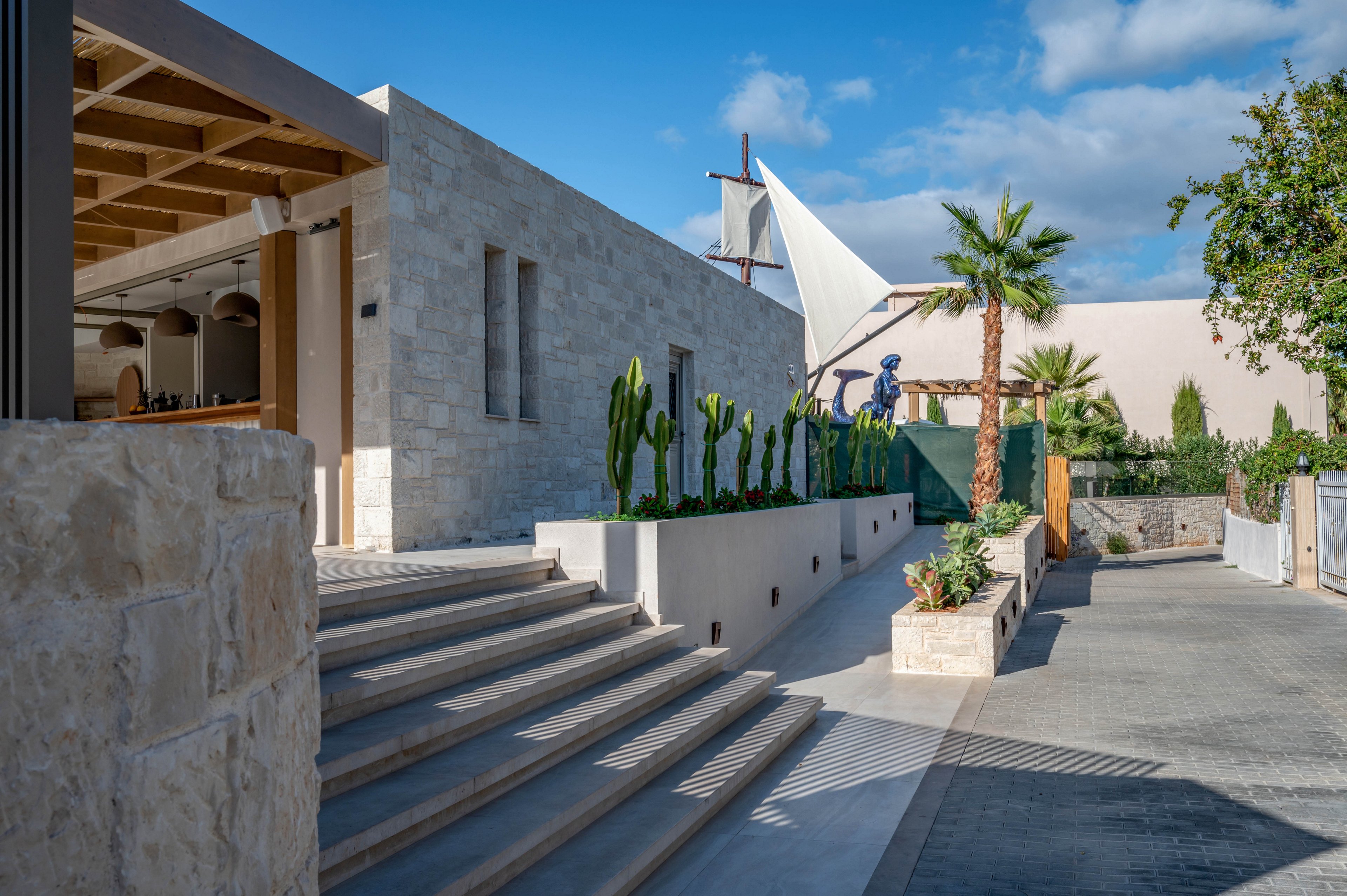 A contemporary stone structure is featured with wide steps leading up to an open entrance. The setting is outdoors, with a row of tall cacti planted along the wall and a palm tree visible in the courtyard. The sky is clear and blue, creating a bright and inviting mood. Architectural details include clean lines and a shaded patio area, with no visible text or numbers.
