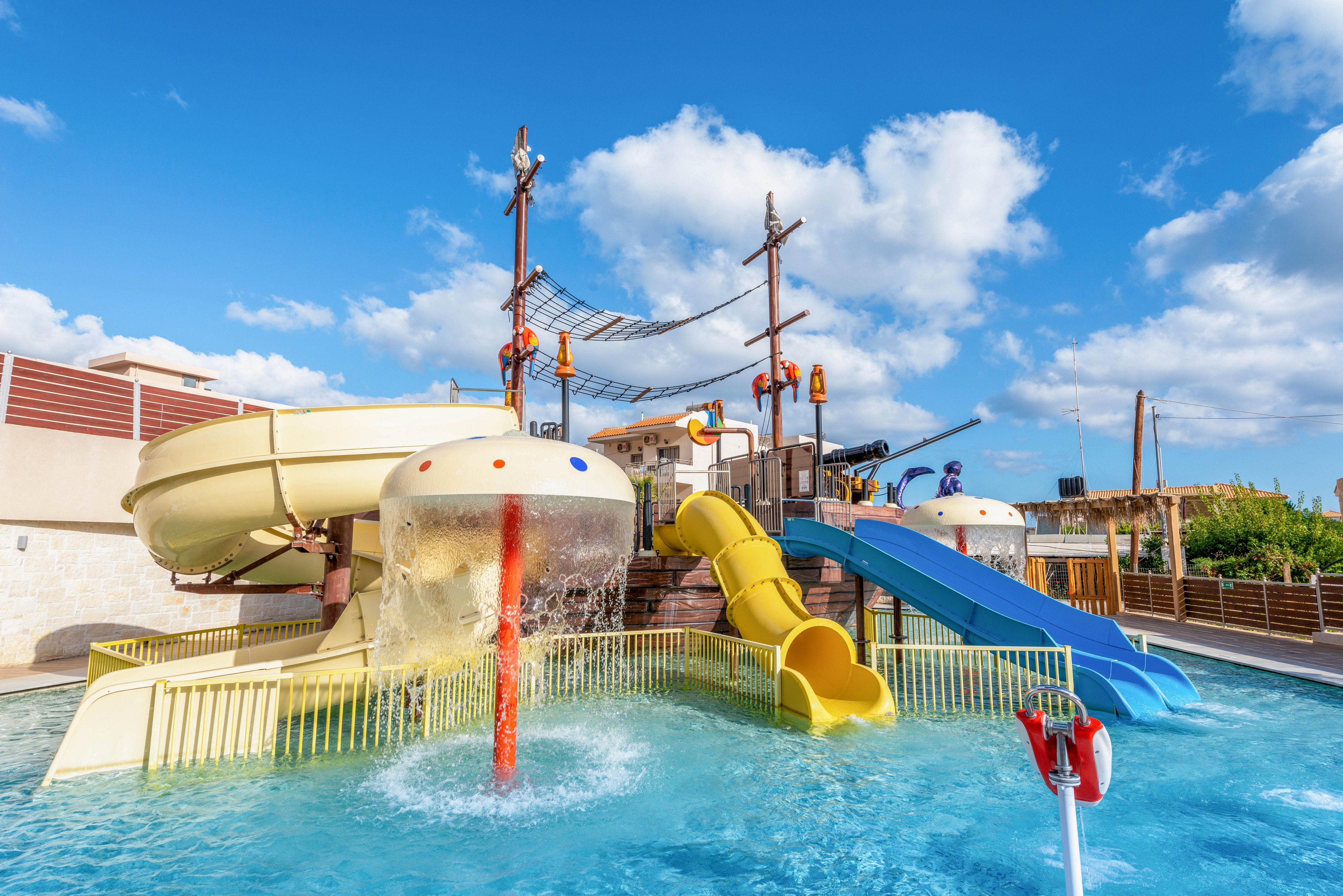 The image features a vibrant outdoor water park with multiple slides, including yellow, blue, and beige tubes, leading into a shallow pool. The setting is sunny with a bright blue sky and scattered clouds, creating a cheerful and inviting atmosphere. No people are visible, and the area is surrounded by fencing and some buildings. The water park equipment stands out with playful colors and dynamic shapes.