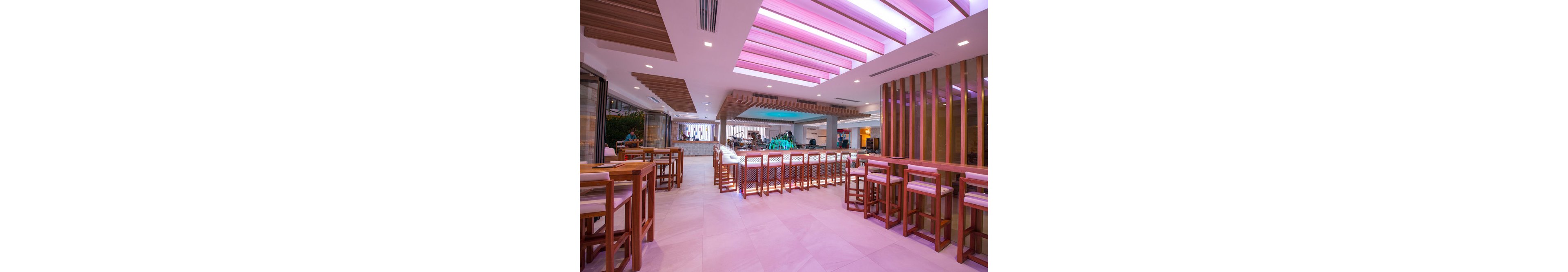The image shows a contemporary lounge bar featuring wooden high tables and chairs. The setting is indoors with a spacious layout, illuminated by pink and purple ceiling lights. The bar area is visible in the background, with a clean and stylish design. No visible text or numeric values are present in the image.