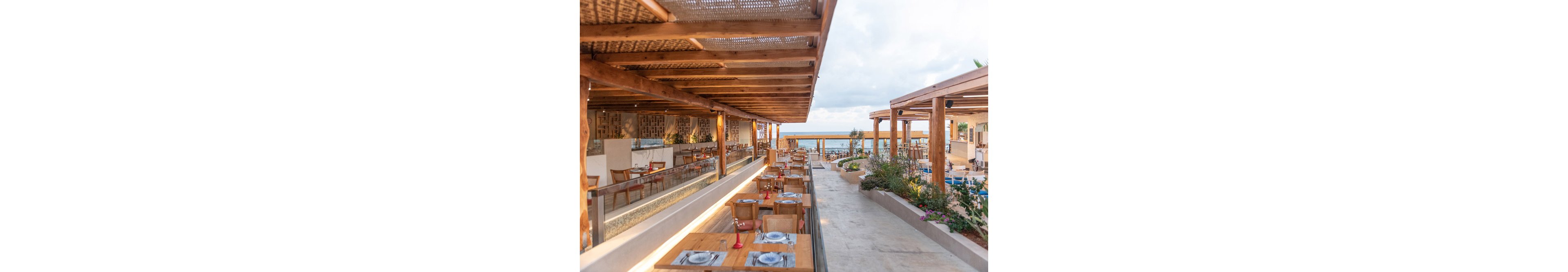 A spacious open-air restaurant features wooden tables and chairs under a pergola roof, overlooking the ocean. The setting is bright and airy, with natural light and coastal landscaping enhancing the relaxed atmosphere. Table settings are neatly arranged, and the overall palette is warm with natural wood tones and touches of greenery. The scene is tranquil, ideal for casual dining by the sea.