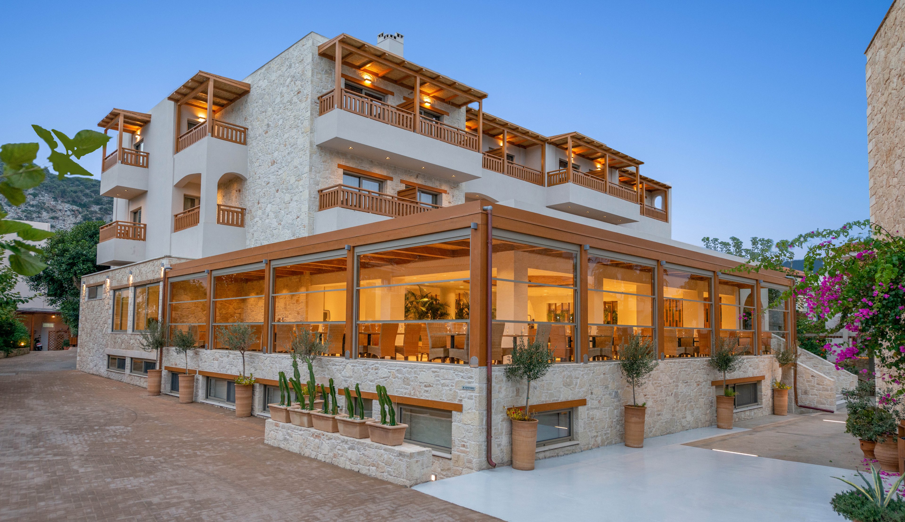 A contemporary multi-story hotel building features stone walls and wooden balconies, illuminated warmly at dusk. The ground floor showcases a spacious glass-enclosed terrace with visible dining tables inside. Potted plants and cacti line the exterior, while the clear sky and surrounding greenery enhance the tranquil atmosphere.