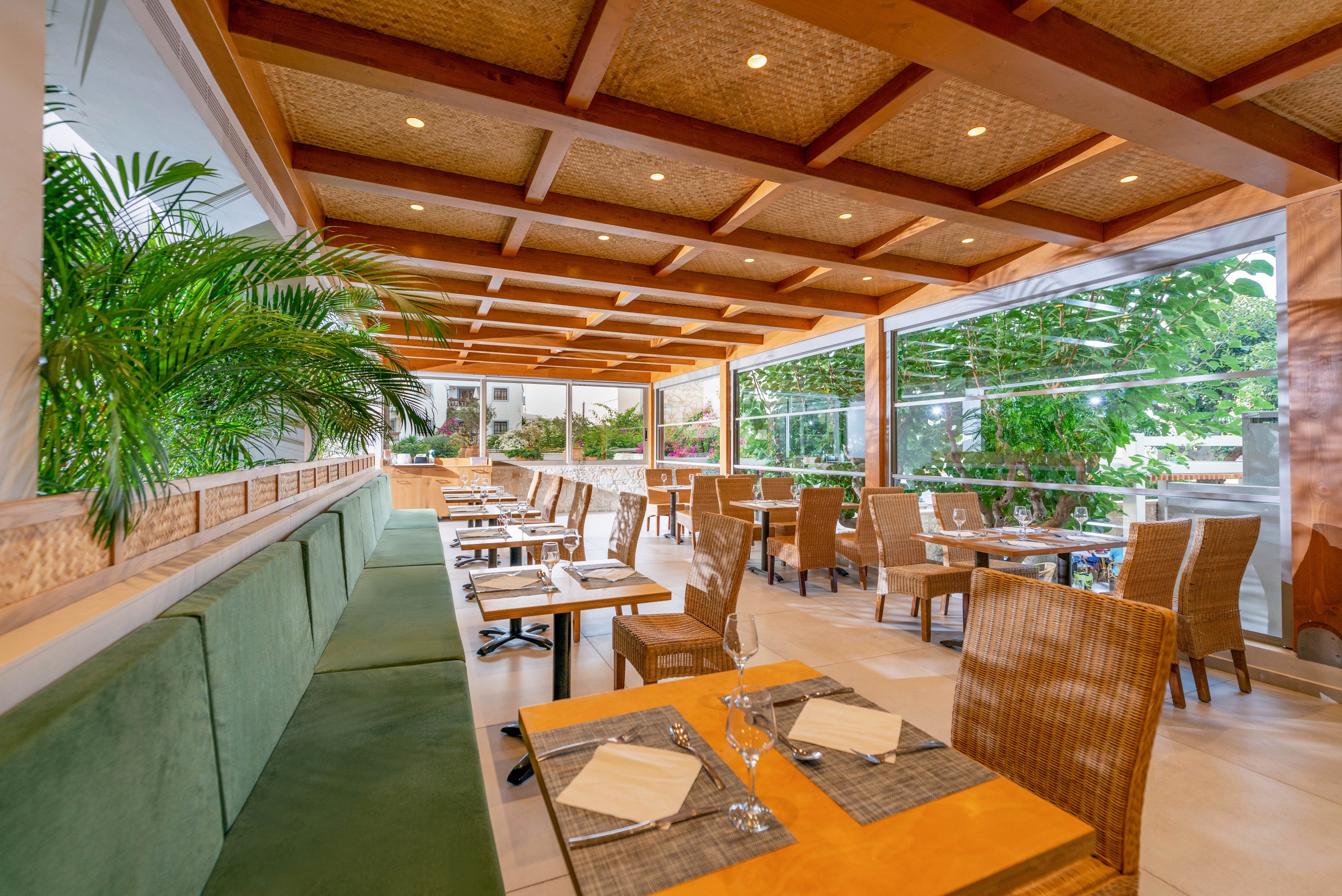 A contemporary restaurant interior features wooden tables and wicker chairs arranged neatly in an open-air setting. Large windows provide views of lush greenery outside, while the ceiling showcases exposed wooden beams. The atmosphere is bright and inviting, with neutral tones and green accents from plants and upholstery.