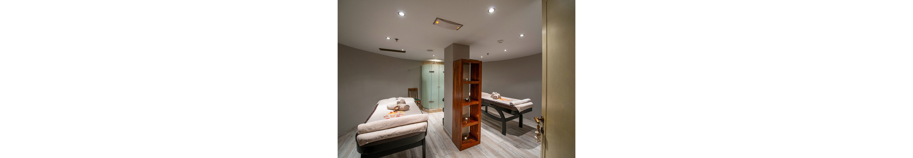 The image shows a contemporary spa treatment room featuring two massage tables with neatly folded towels and flower decorations. The setting is calm and inviting, with soft lighting and a wooden shelving unit dividing the space. A glass shower enclosure and a wooden chair are visible in the background, enhancing the tranquil atmosphere. The overall palette is neutral, with beige and brown tones contributing to a relaxing mood.