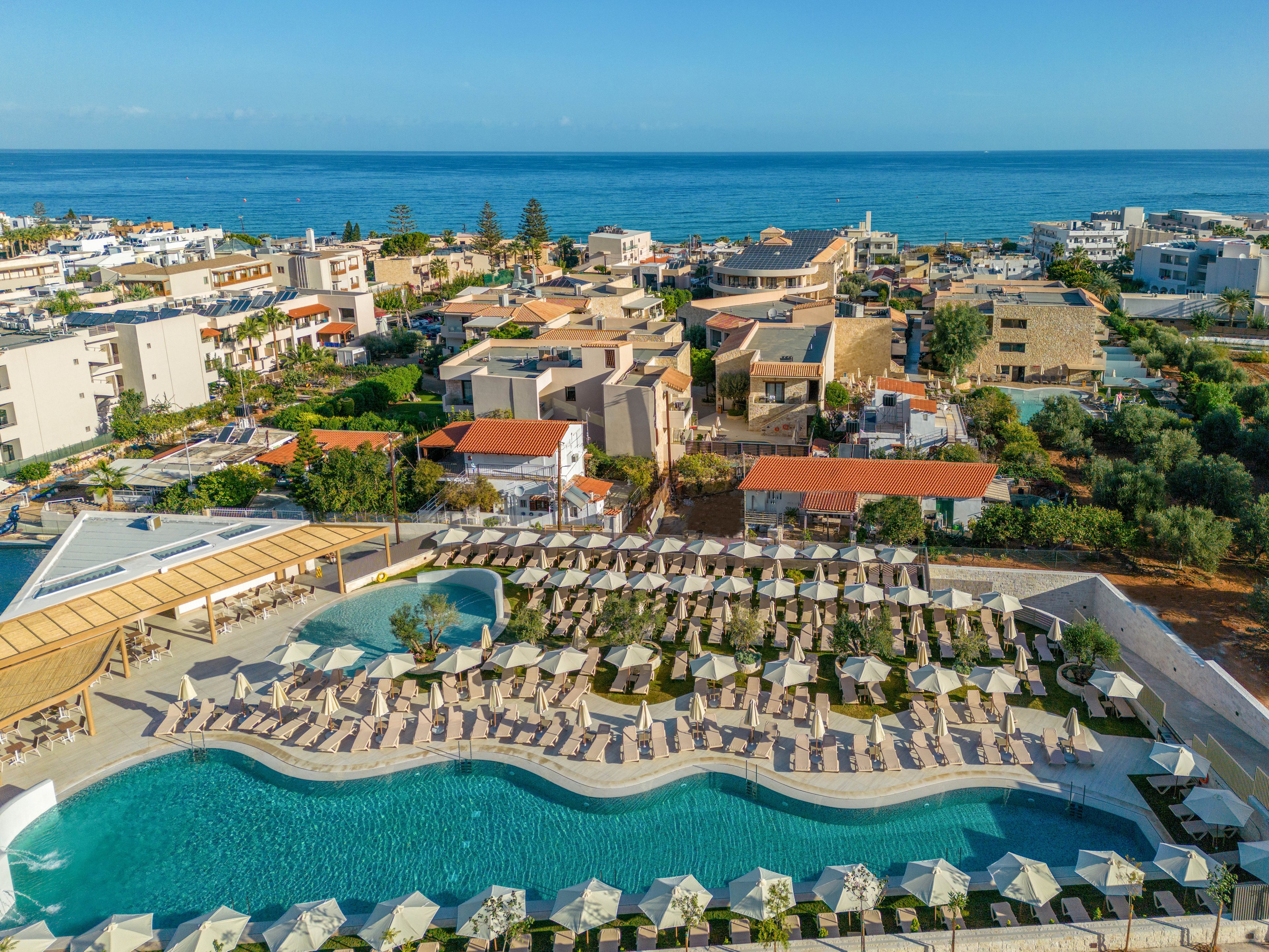 The image shows a large resort complex featuring a curving swimming pool surrounded by numerous sun loungers and umbrellas. The setting is coastal, with the sea visible in the background and a mix of residential and hotel buildings nearby. The scene is bright and sunny, evoking a relaxed vacation mood with a palette of blues, greens, and neutrals. No visible text or numbers are present in the image.