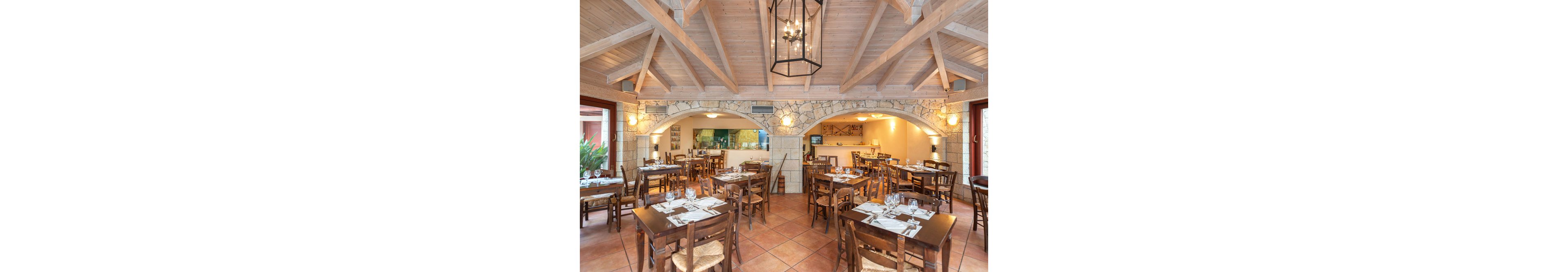 A spacious restaurant dining area features wooden tables and chairs arranged neatly under a vaulted ceiling with exposed beams. The setting is warm and inviting, with stone archways and large windows allowing natural light to fill the space. Table settings include glassware, plates, and napkins, creating a ready-to-serve atmosphere. The color palette is earthy with natural wood tones and terracotta floor tiles. No visible text or numbers are present in the image.