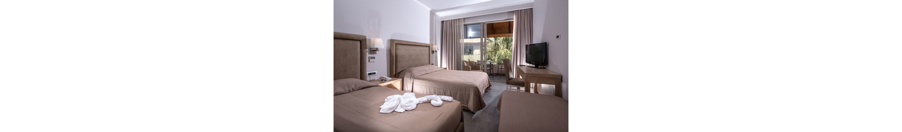 A contemporary hotel room features two twin beds and one single bed, all dressed in neutral brown bedding. The room is well-lit with wall-mounted lamps and natural light from a large window leading to a balcony with outdoor seating and palm trees visible. A towel arrangement resembling a swan is placed on one bed, and a flat-screen television sits atop a wooden desk. The overall palette is soft and muted, creating a calm and inviting atmosphere.