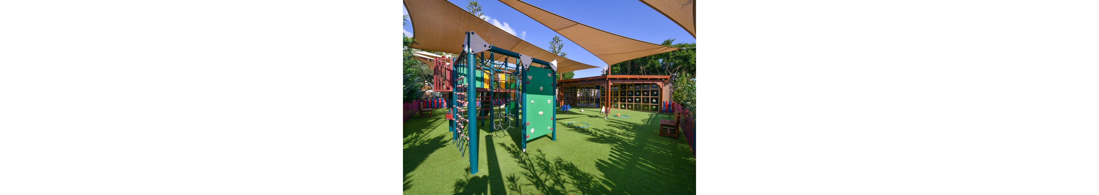 A vibrant outdoor playground features a green climbing structure with ropes and panels, set on artificial grass. The area is shaded by large fabric canopies and surrounded by colorful fencing. Play equipment and toys are scattered around, with a building in the background displaying a wall of cubby holes. The scene is bright and inviting, ideal for children’s activities.
