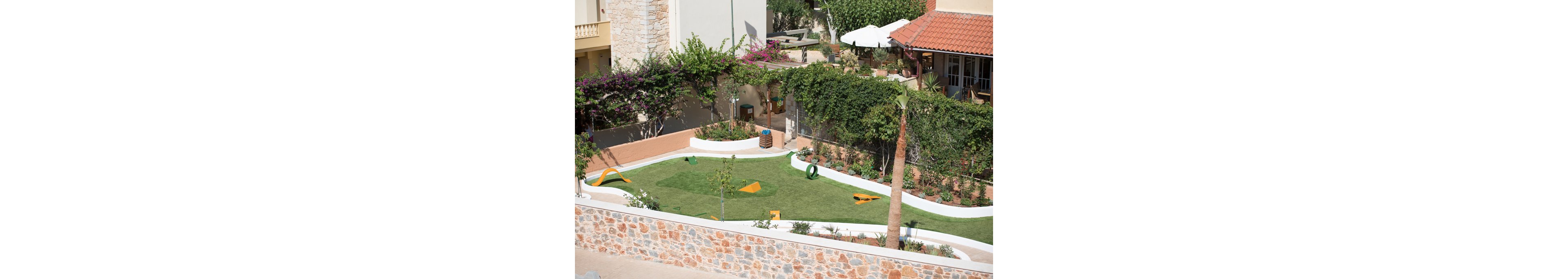 The image shows a small, enclosed dog agility park featuring various yellow and green obstacles on a well-maintained grassy lawn. The setting is outdoors, surrounded by stone walls and lush greenery, with residential buildings and patio furniture visible in the background. The park includes ramps, tunnels, and jumps, creating a playful and inviting atmosphere. No people or visible text are present in the scene.