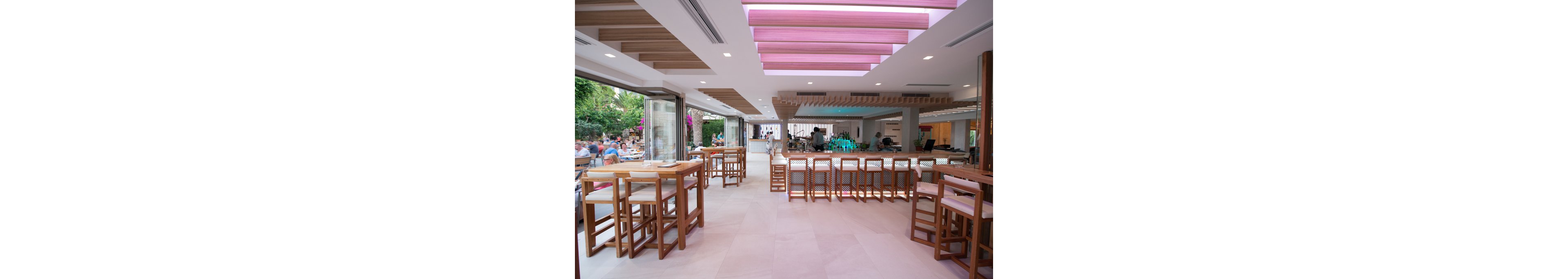 A spacious, contemporary restaurant features an open-air layout with wooden tables and high chairs. The bar area is centrally located, illuminated by soft pink and white ceiling lights. Large windows open to an outdoor dining area with greenery and patrons enjoying their meals. The interior design emphasizes clean lines, natural light, and a relaxed, inviting atmosphere.