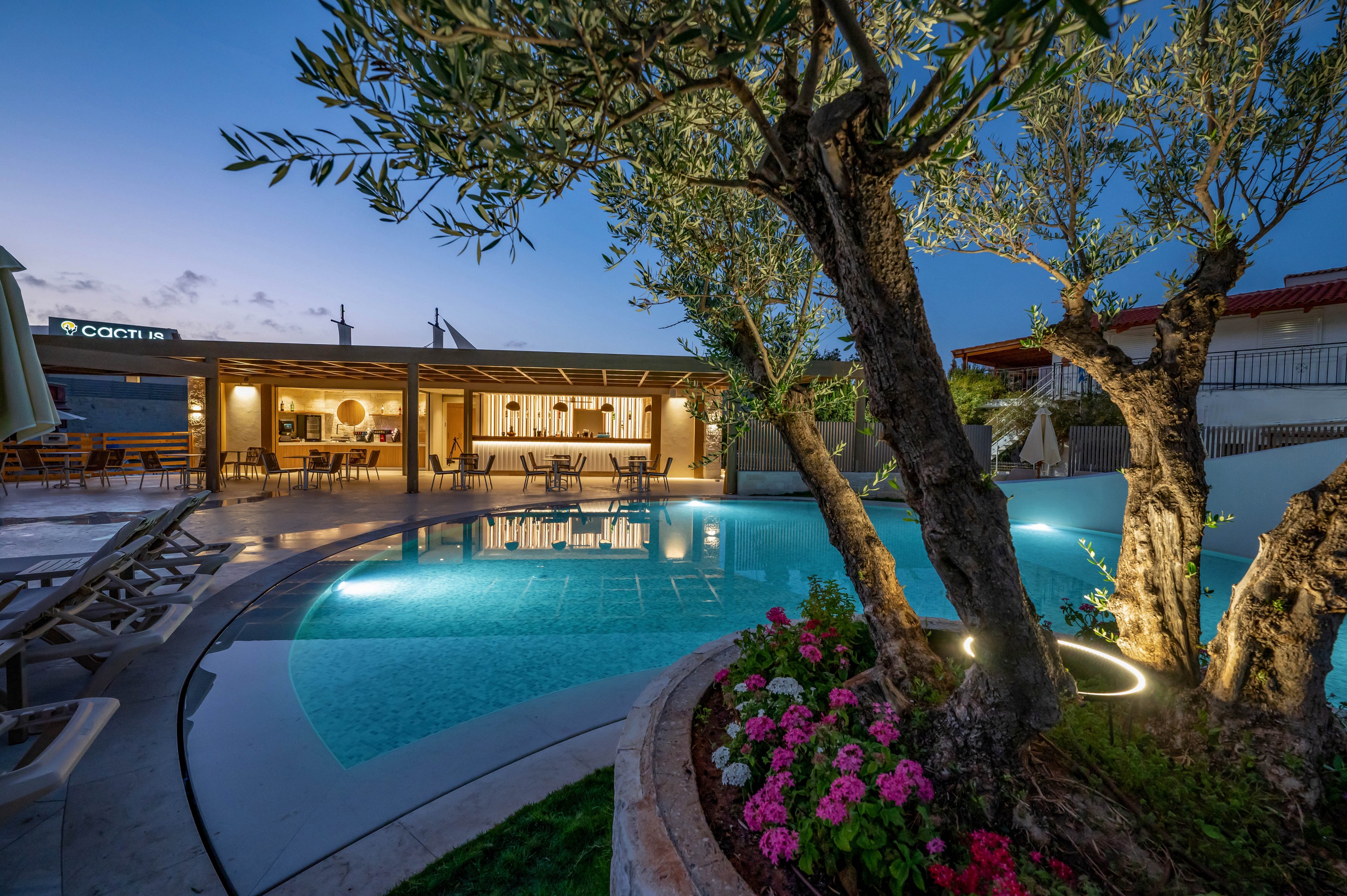 A contemporary swimming pool is featured in a landscaped outdoor setting at dusk. The scene includes illuminated trees, vibrant flower beds, and a stylish poolside bar with seating. Reflections from the pool and soft ambient lighting create a tranquil atmosphere. The visible text 'COCHI' appears on a building in the background, adding a distinct element to the scene.