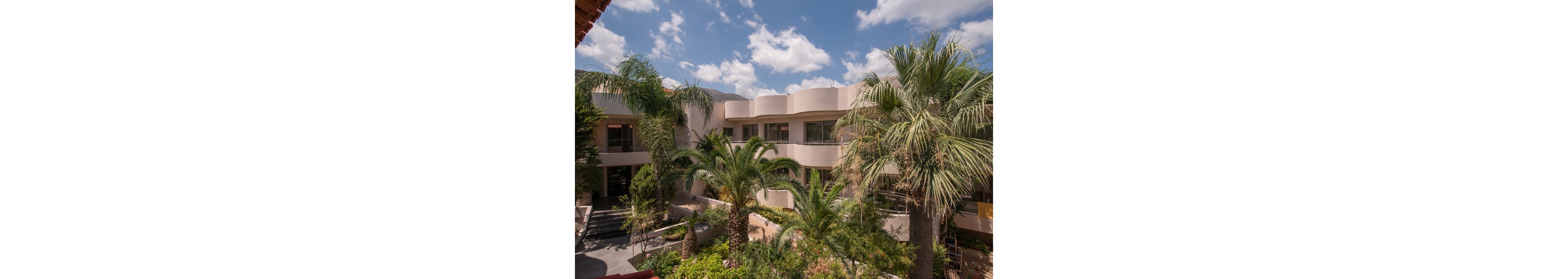The main subject is a contemporary, multi-story building with curved balconies and large windows. The setting features lush landscaping with several palm trees and tropical plants, creating a vibrant and inviting atmosphere. The sky is bright blue with scattered clouds, enhancing the warm and sunny mood. No visible text or numeric values are present in the image.