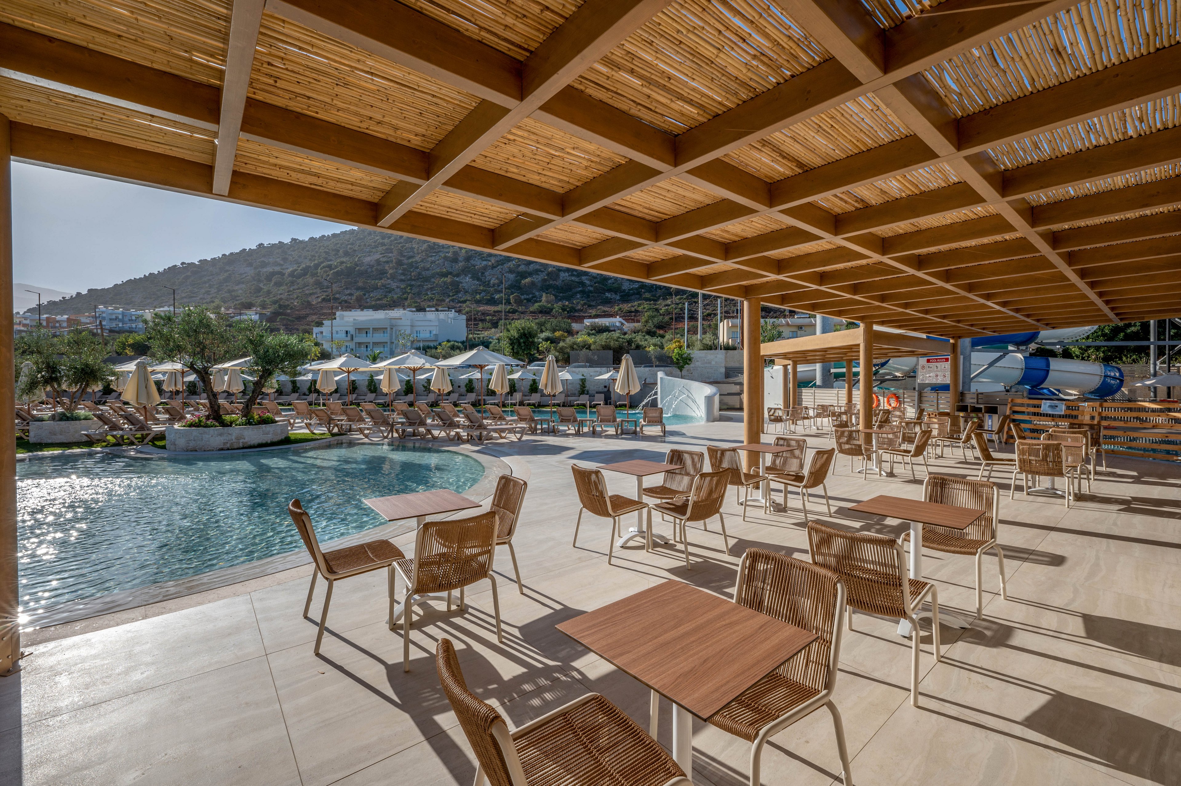 The image shows a spacious outdoor seating area with wooden tables and chairs under a shaded pergola. In the background, there is a swimming pool surrounded by numerous sun loungers and umbrellas, set against a hillside with buildings. The scene is bright and sunny, featuring neutral tones and a relaxed vacation atmosphere. No visible text or numeric values are present in the image.